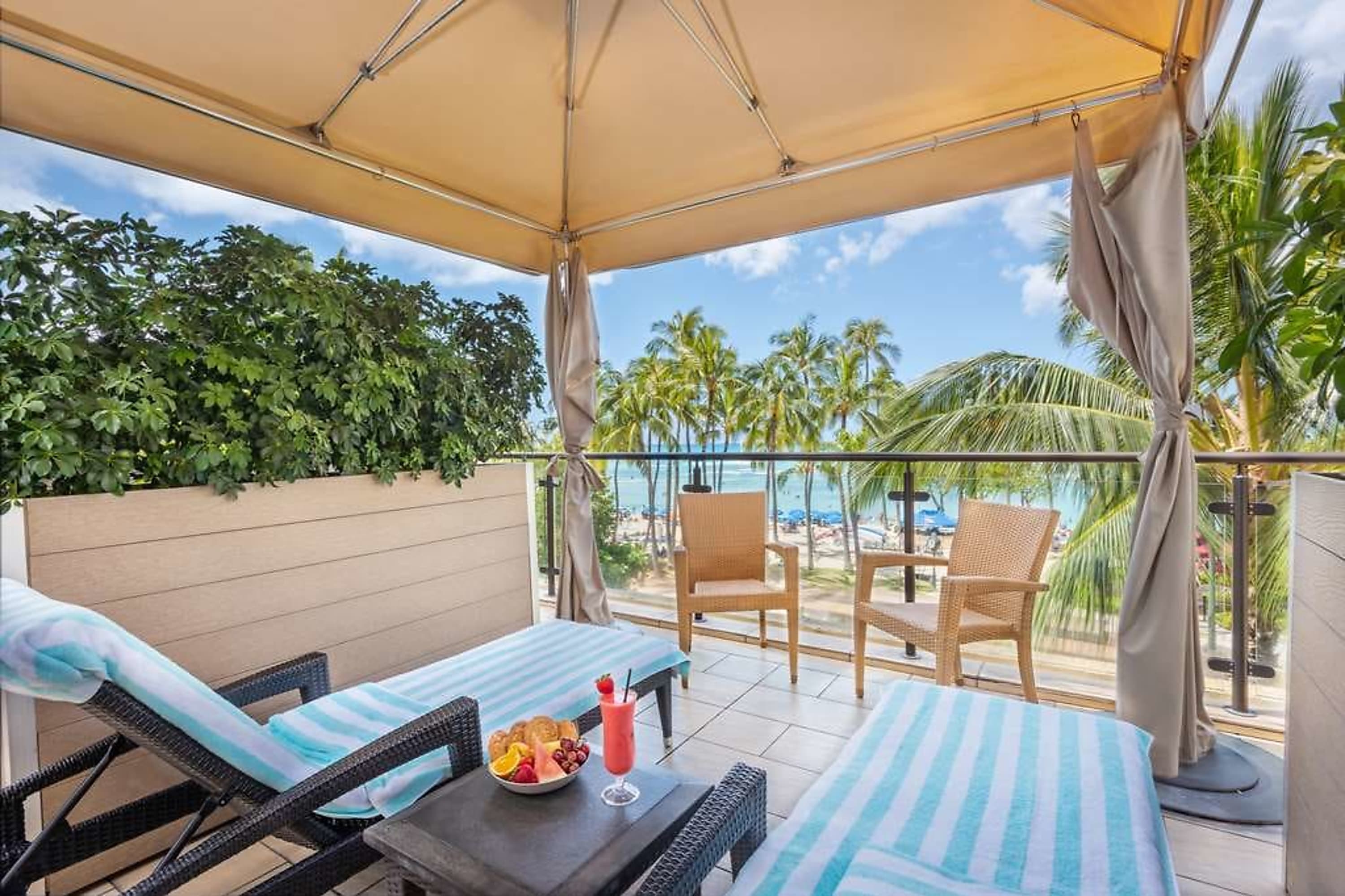 Two loungers and small table with a fruit bowl under an umbrella on a balcony with beach views