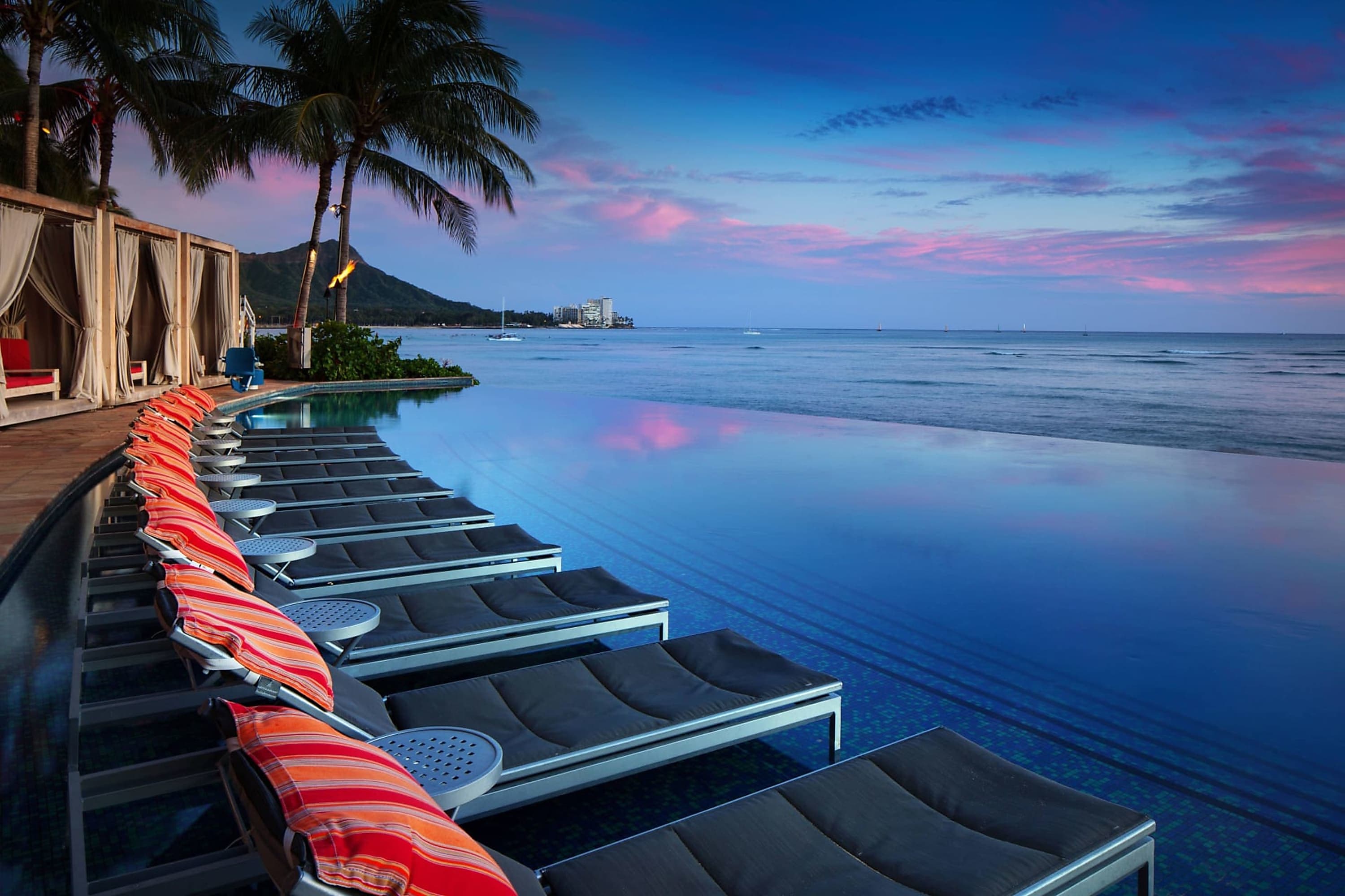 Pool chairs lined along one edge of a lengthy infinity pool; the other edge looks out over the Pacific