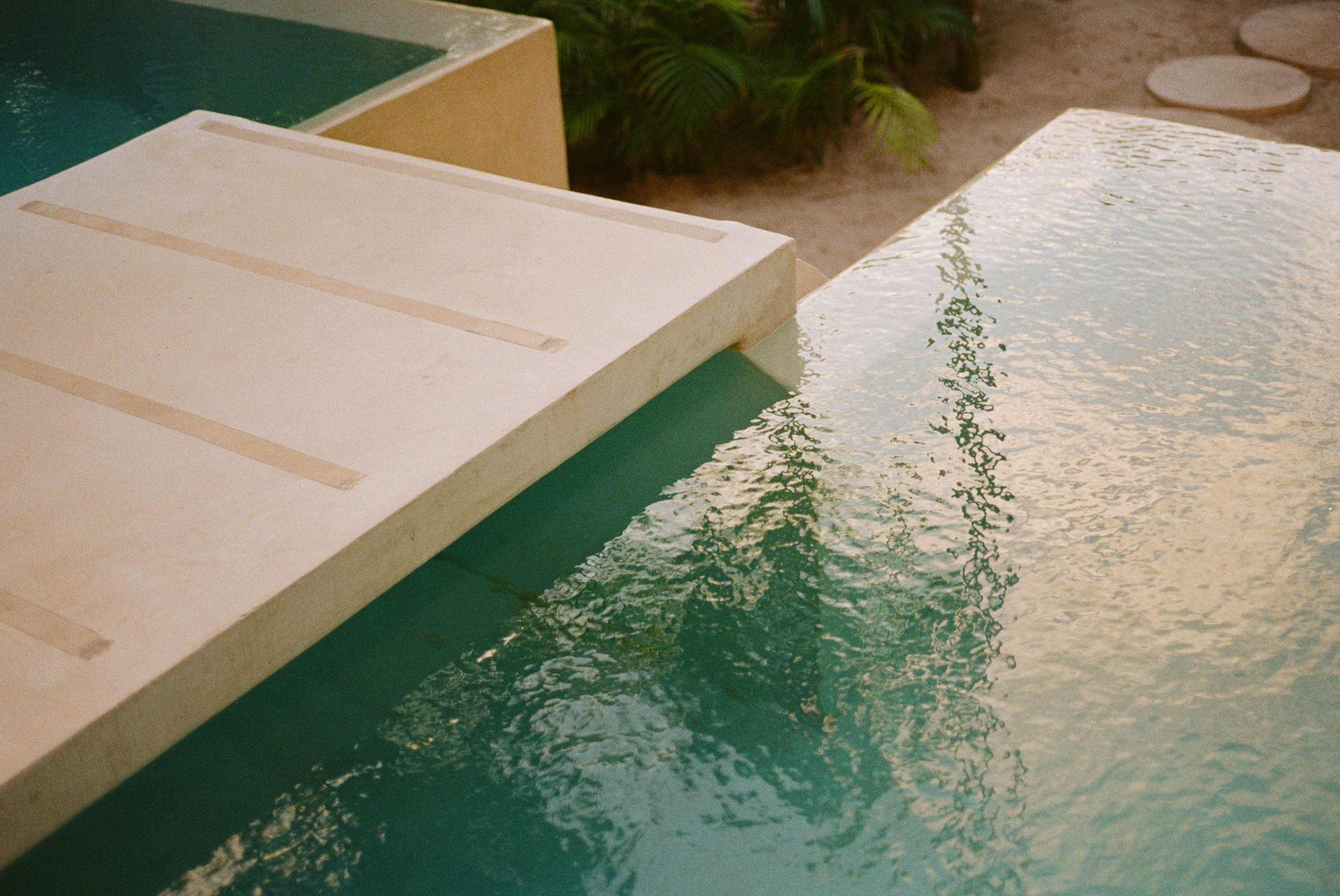 An infinity pool with sand and foliage in the distance.