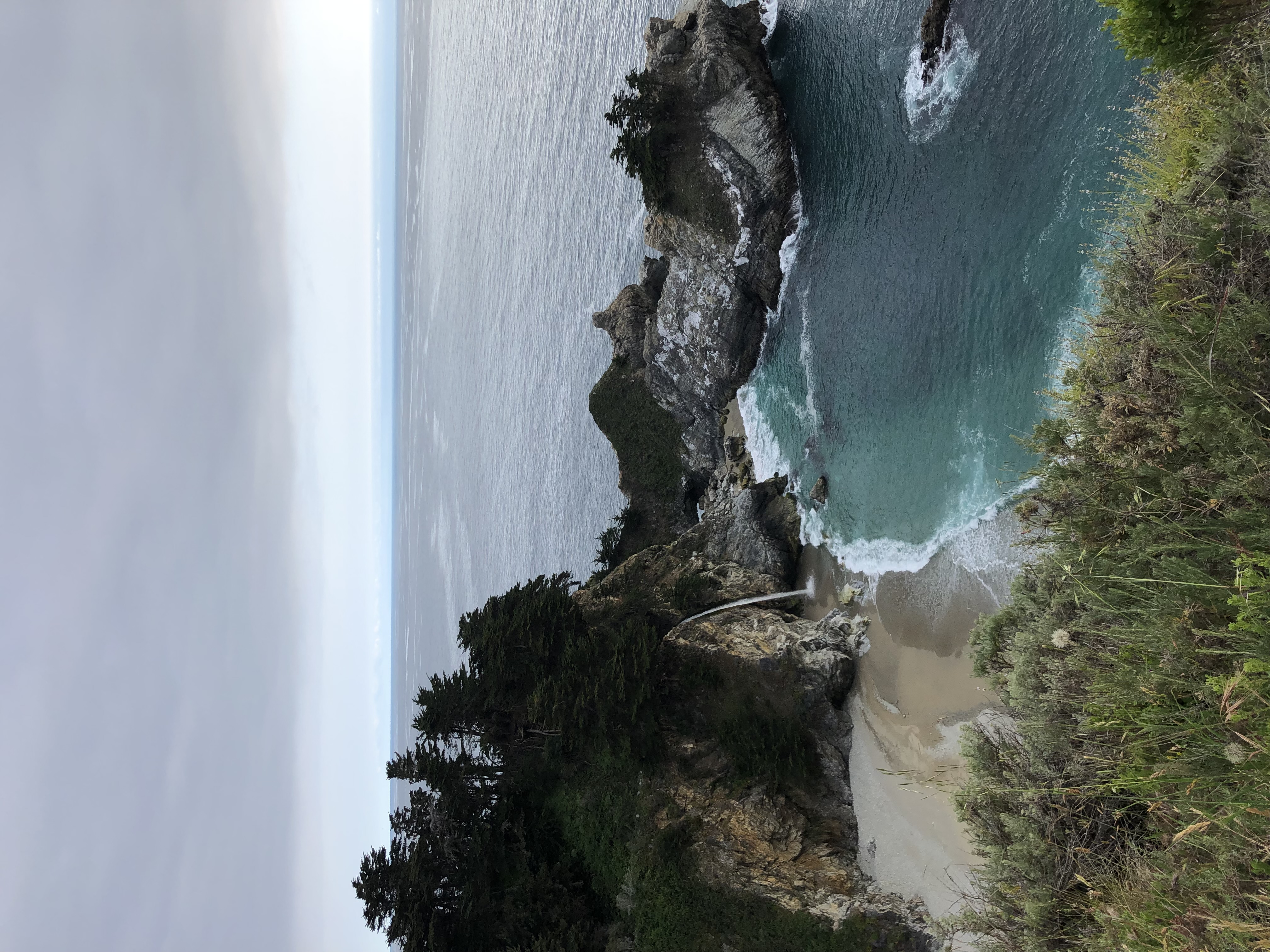 A high up image of a mountain scape with an ocean cove and a shoreline on a cloudy day.