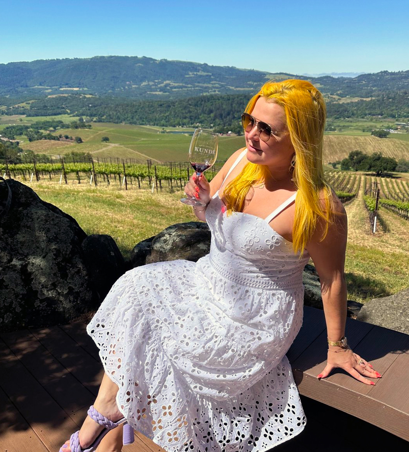 Natalia wearing a long white dress and holding a glass of red wine with a beautiful vineyard behind her