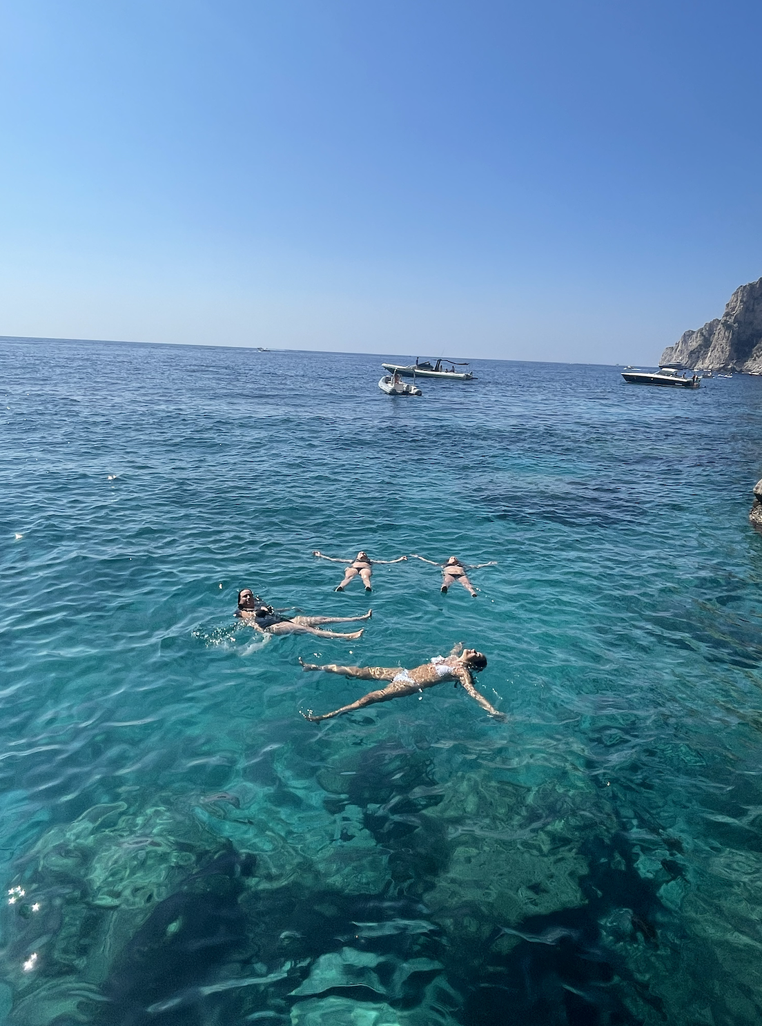 View of Sofia and friends floating on their backs in a crystal clear sea with small boats in the distance
