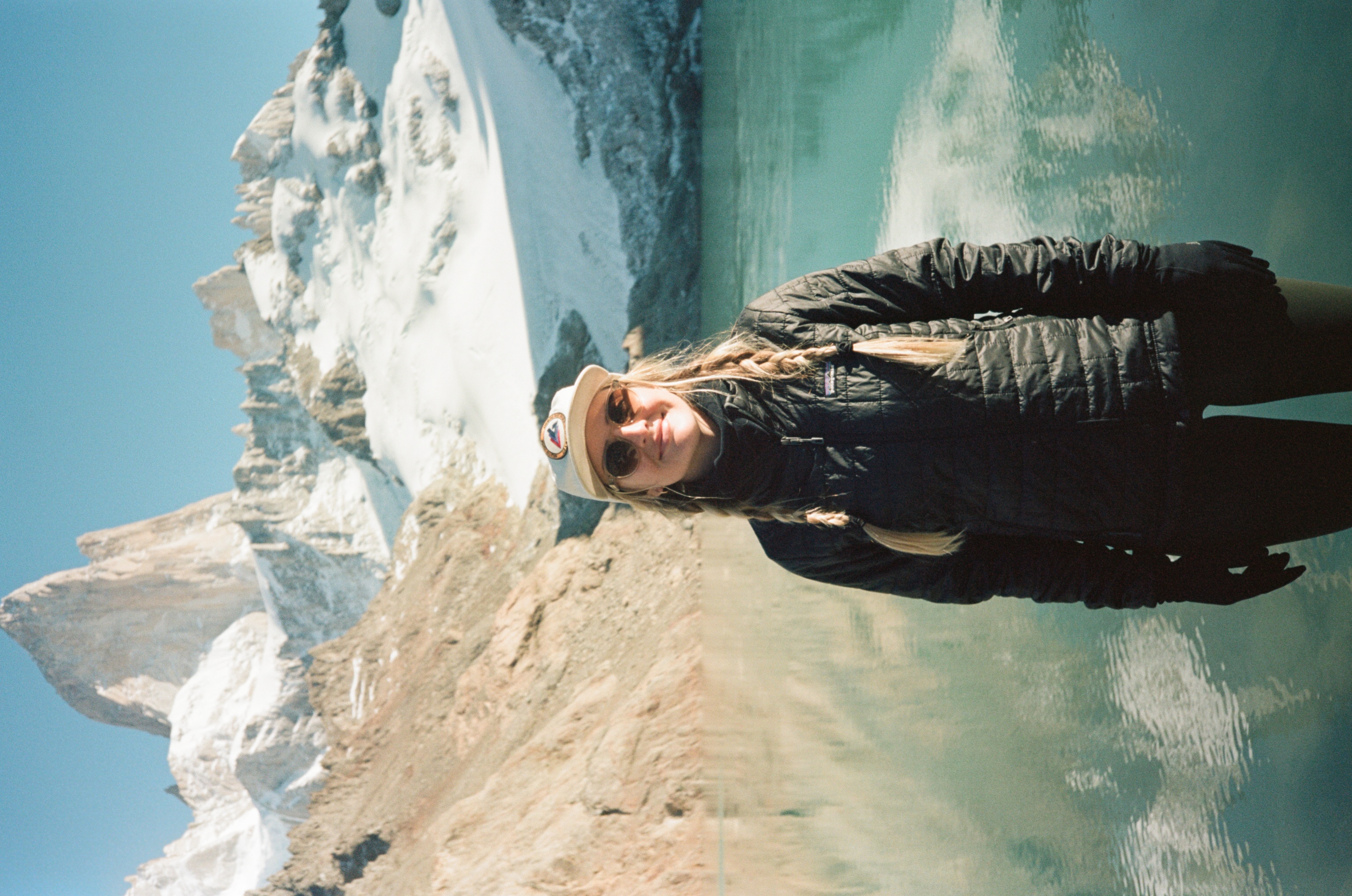 Makenna in a black jacket standing in front of an alpine lake with snowy mountains in the background.