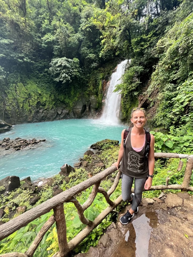 Advisor posing for an image on a wooden deck with a body of water and waterfall in the background with foliage on a sunny day.