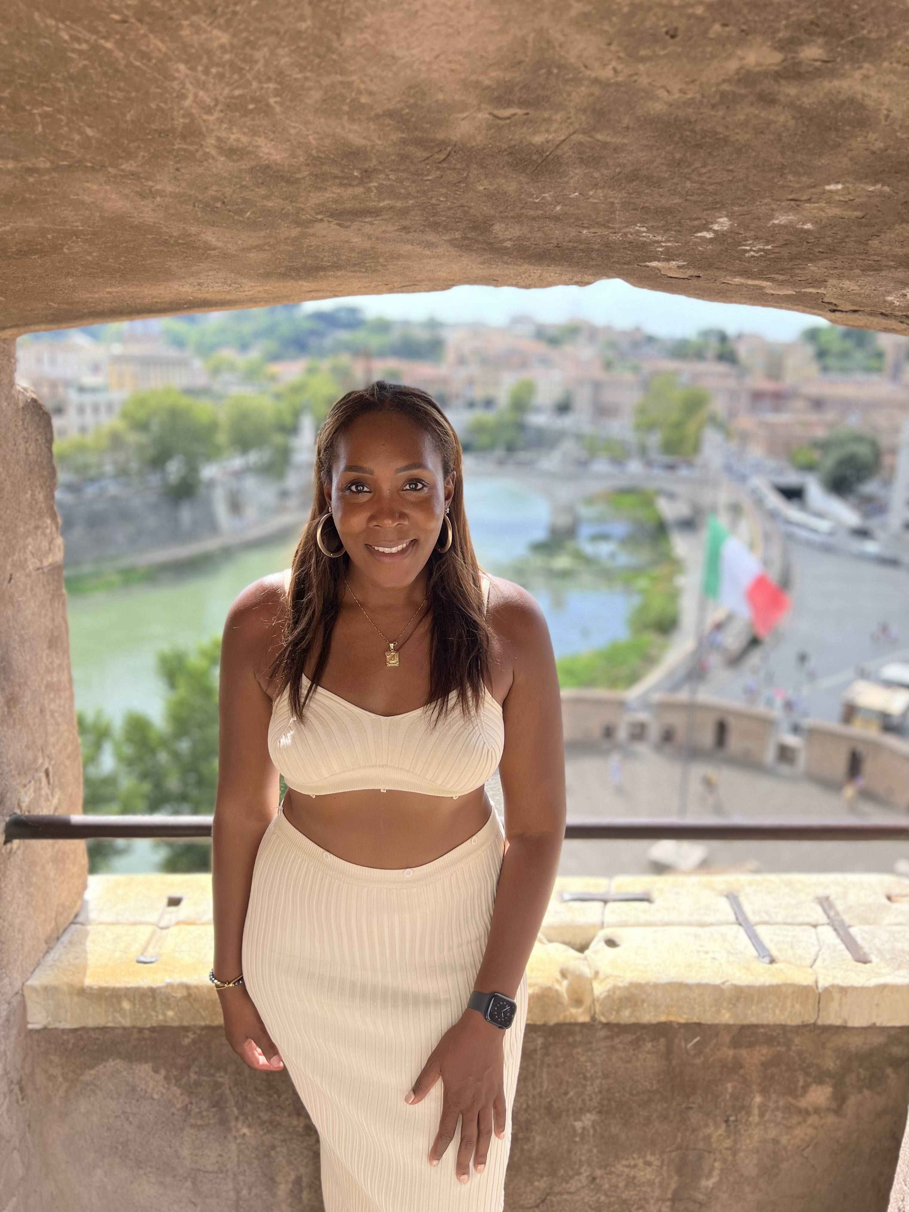 Jessica in a white outfit posing for a photo in a window with a beautiful city view behind her