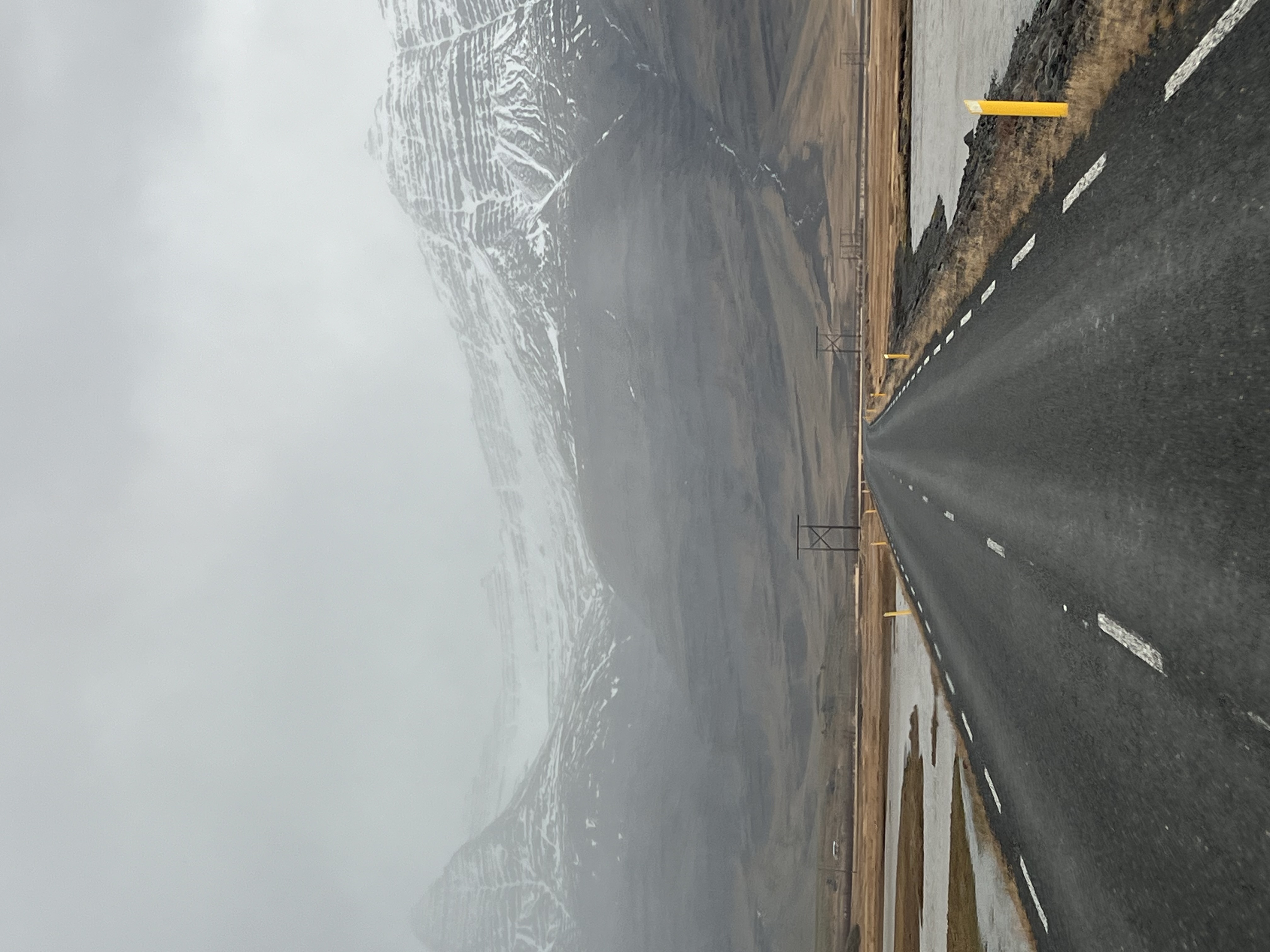 View of an empty road leading towards large snowy mountains slightly covered in clouds