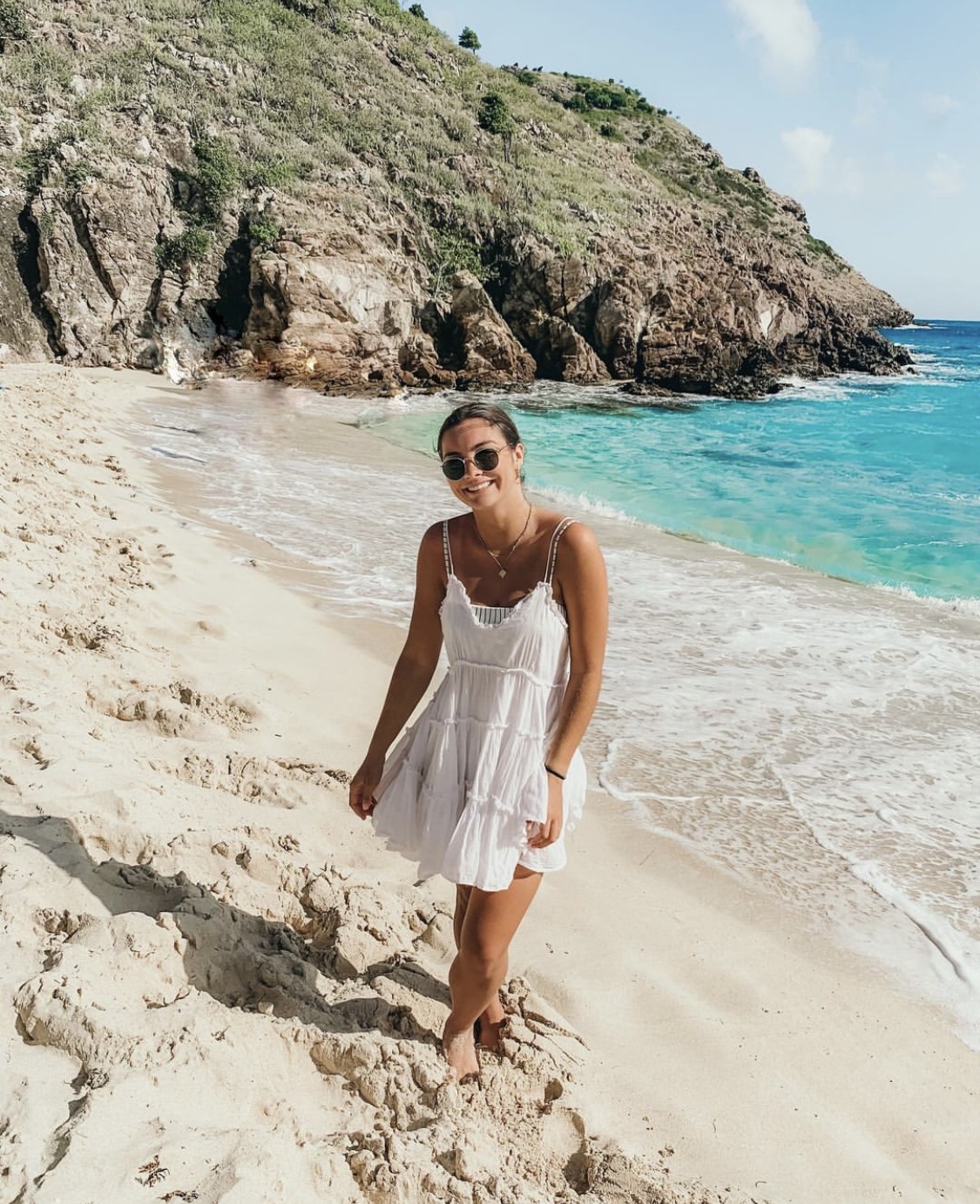 Advisor wearing a white dress on a beach with water and rocky cliffs in the background.
