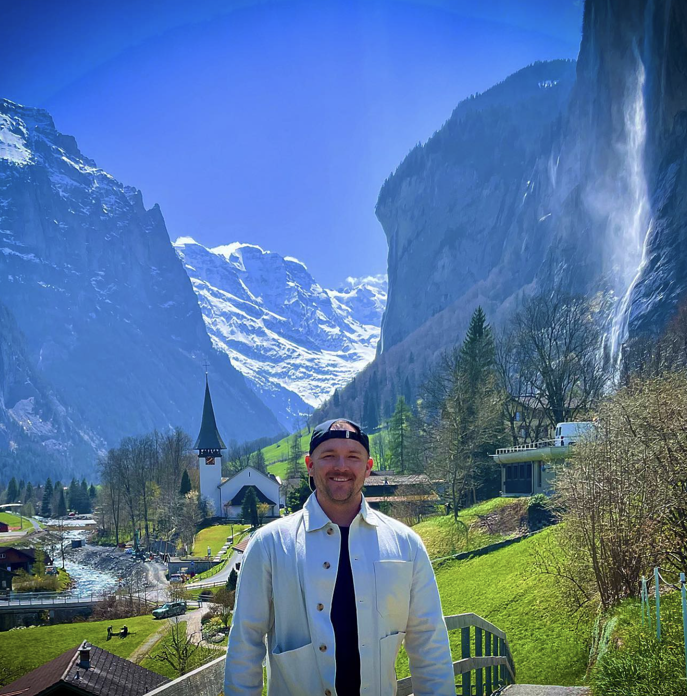Andrew in a white jacket posing in front of a beautiful alpine landscape with snowy mountains on a sunny day
