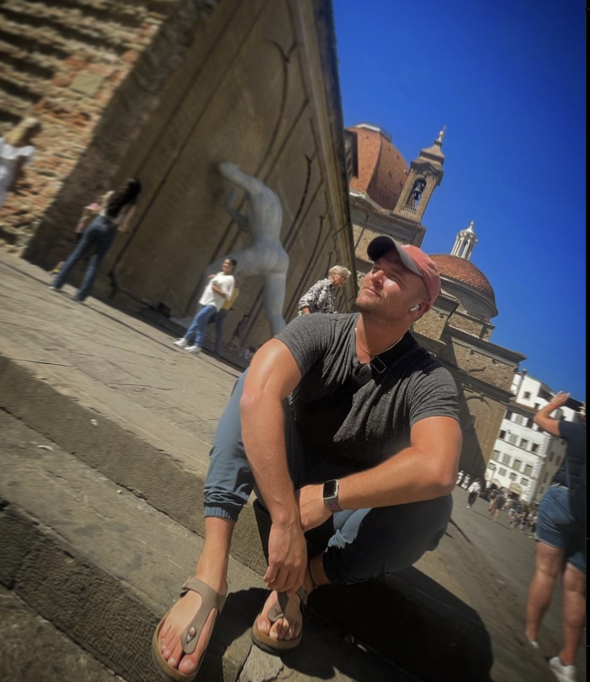 Andrew in a baseball cap and sandals sitting on outdoor steps in front of large monuments on a sunny day