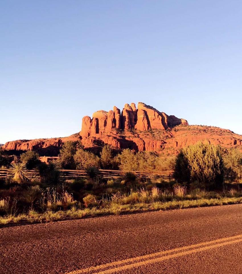 Beautiful view from the road of a large red rock formation surrounded by desert plants under clear blue skies