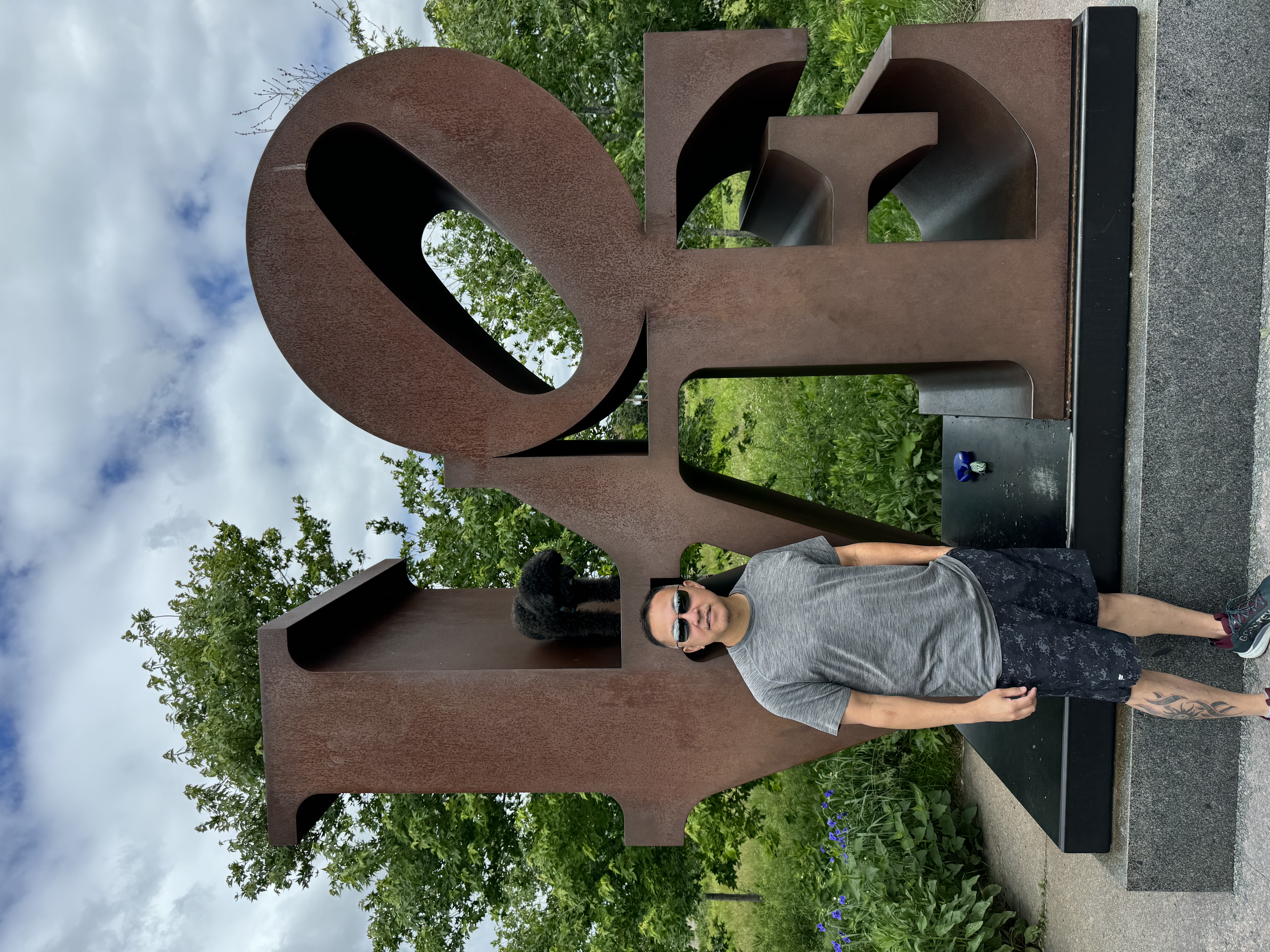 Scott in a grey t-shirt standing in front of a metal sign spelling “LOVE” on a cloudy day
