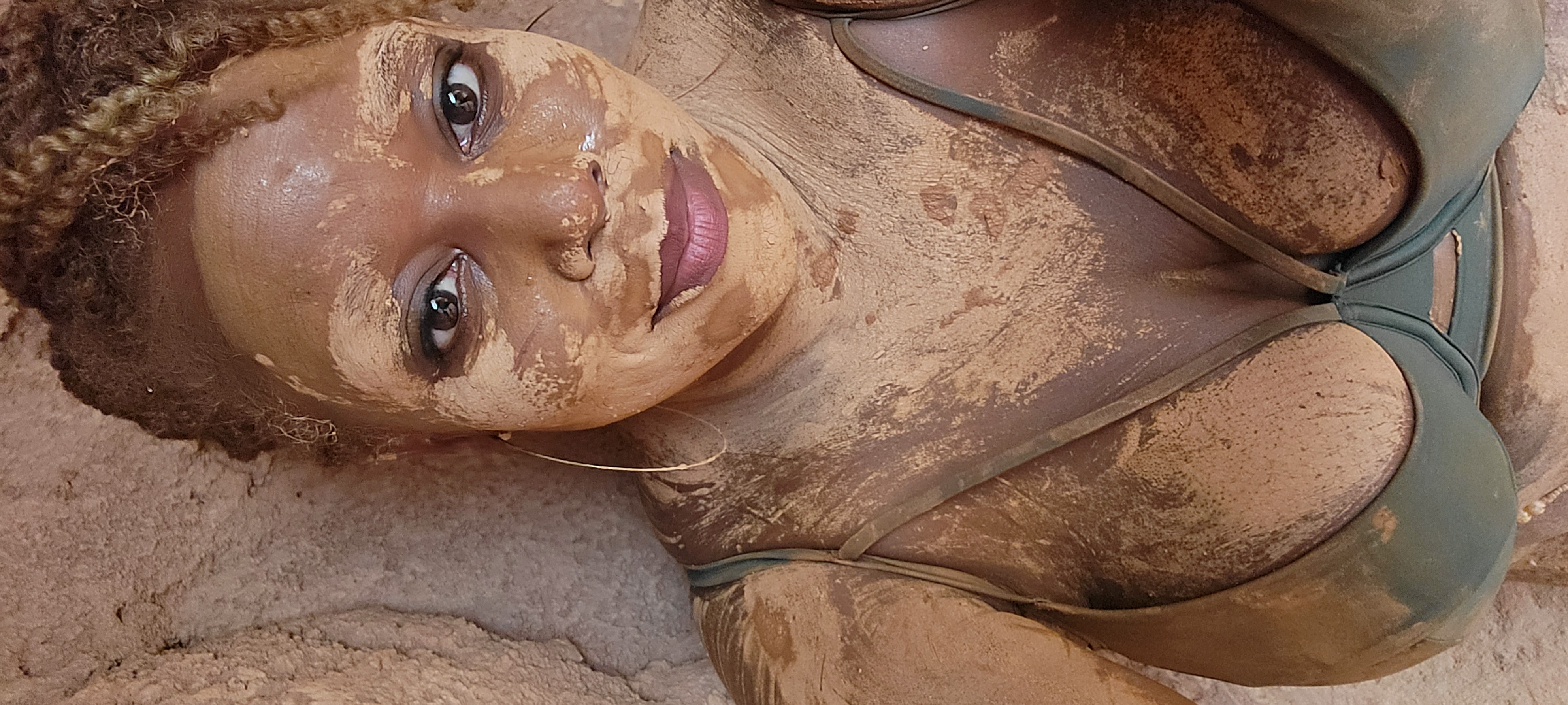 A woman covered in a mud mask relaxing against a stone wall.