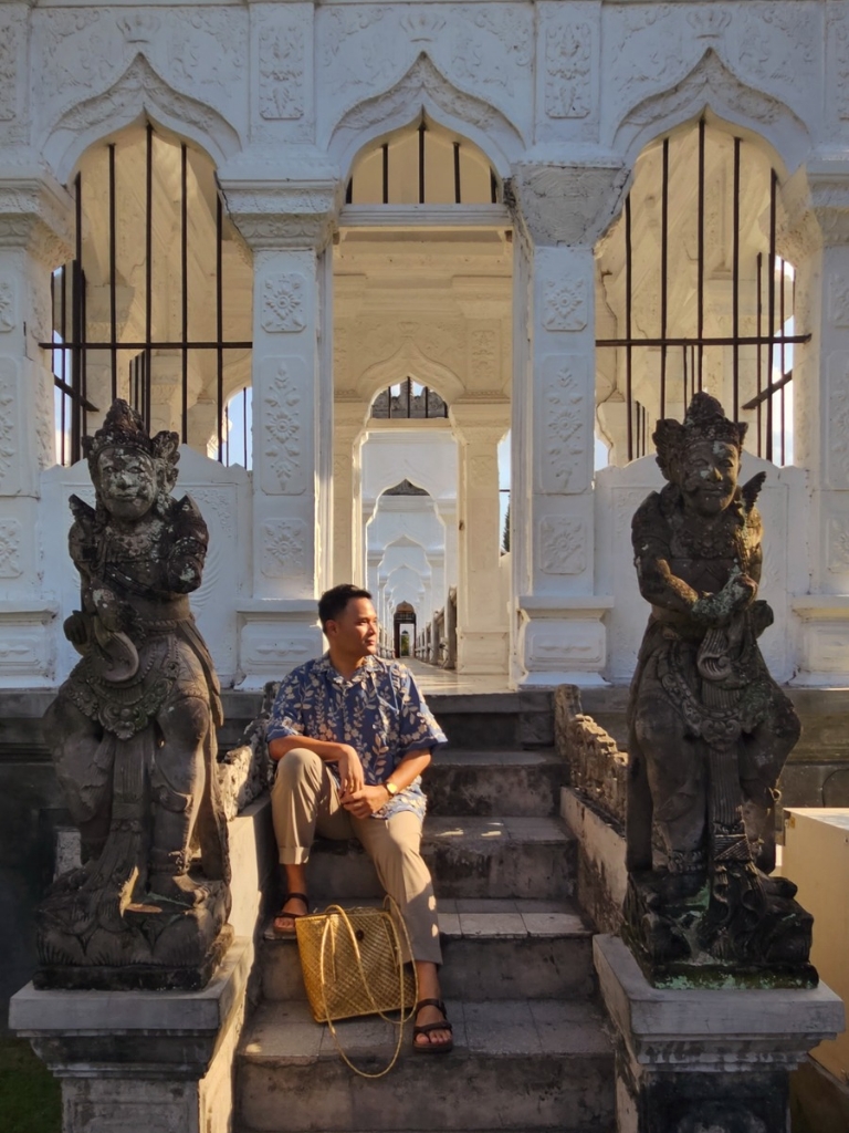 Bima sitting on stone steps in between two old statues of human-like figures, a grand white building behind him
