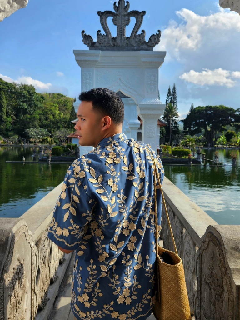 Bima in a blue robe with yellow flowers walking along a narrow wooden bridge over a body of water, a large statue in front of him