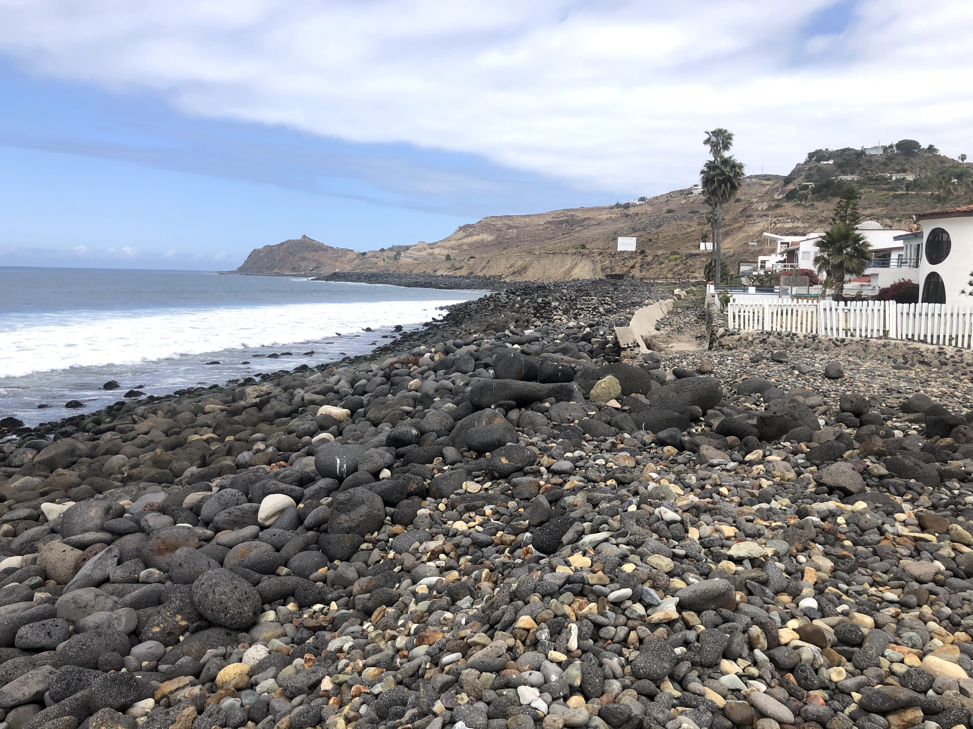 A rocky beach with dark rocks and some white buildings in view.