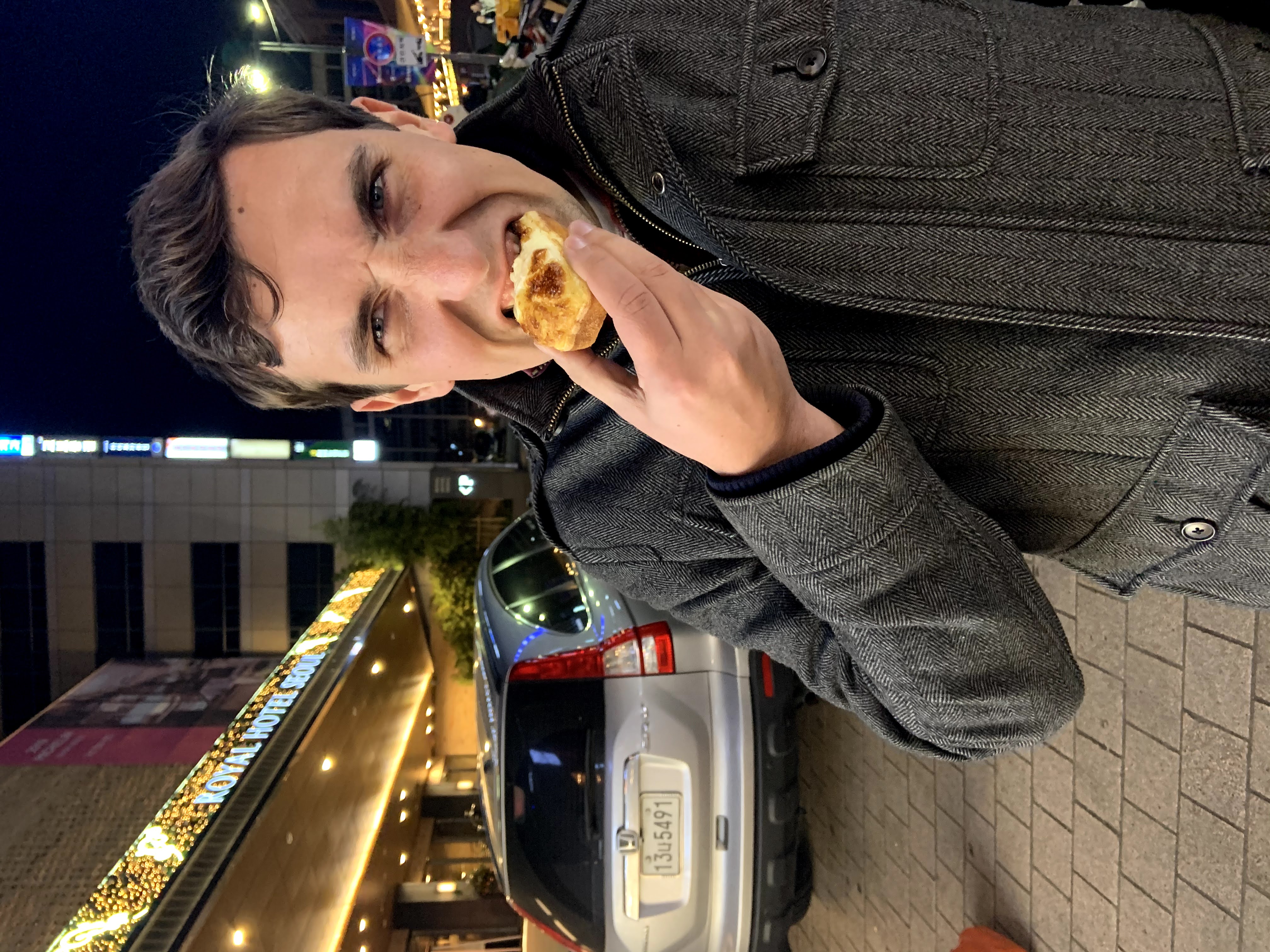 Image of a man in a grey coat biting a pastry on a city street at night