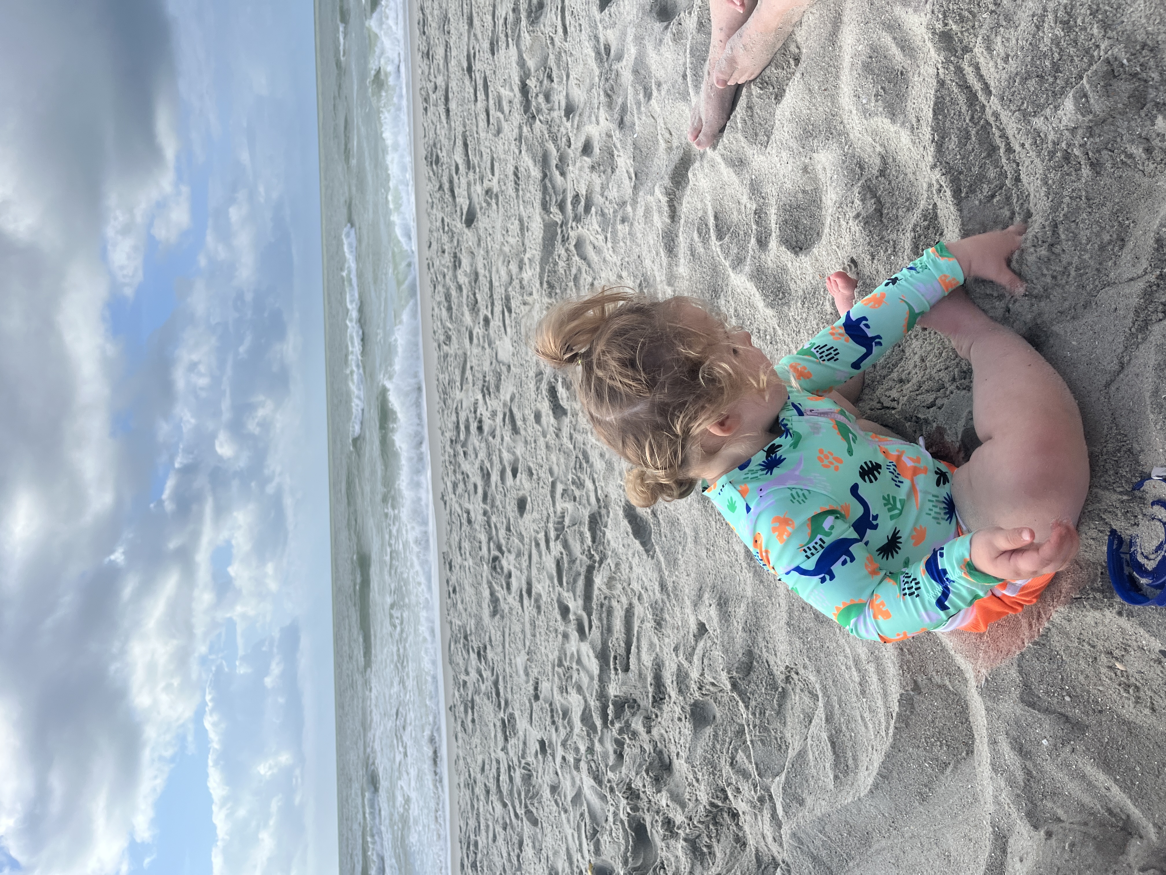 Image of a baby in a blue shirt playing in the sand on the beach with clouds above