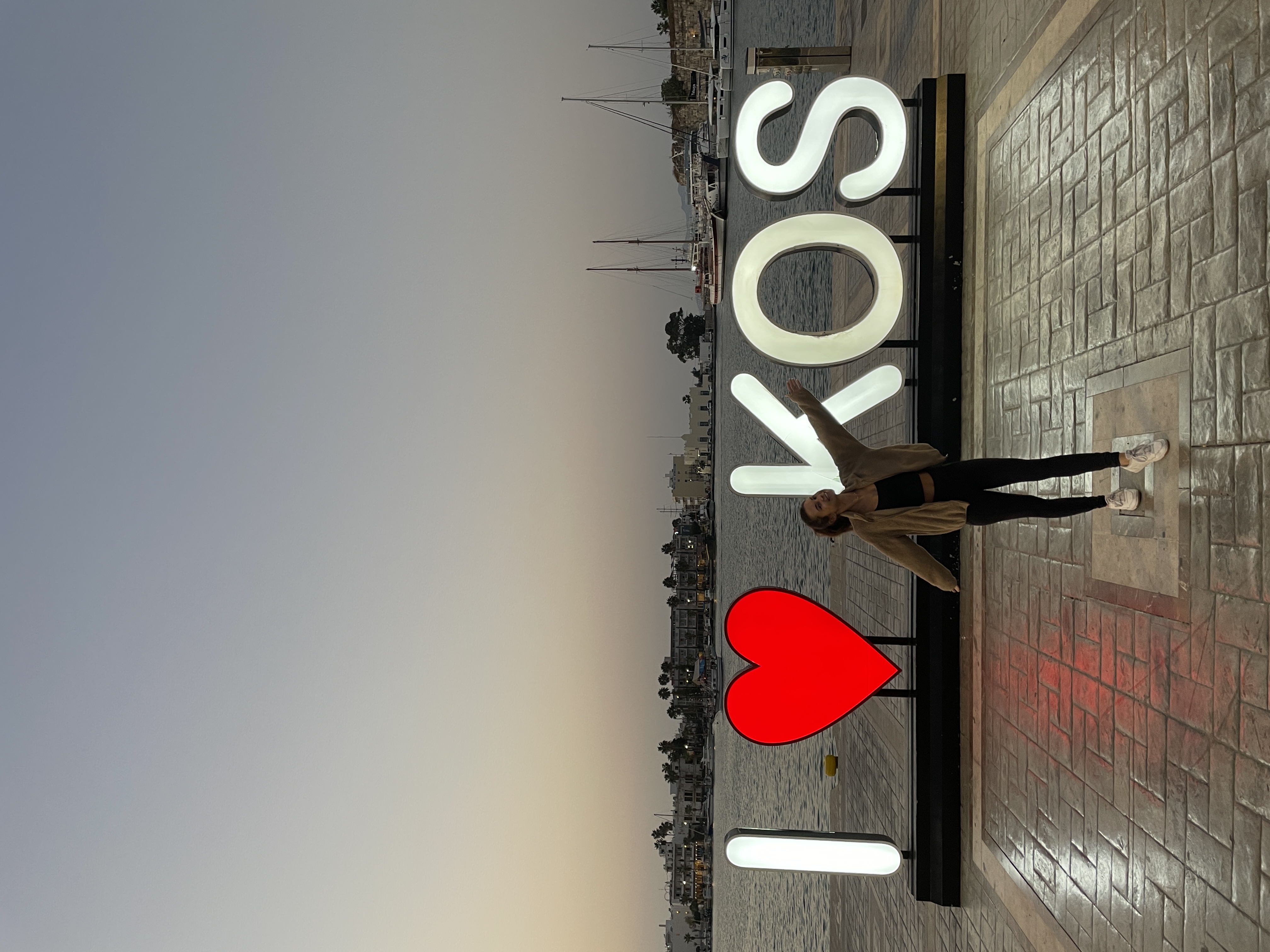 Alayna standing in front of a lit up sign reading “I <3 KOS” at sunset