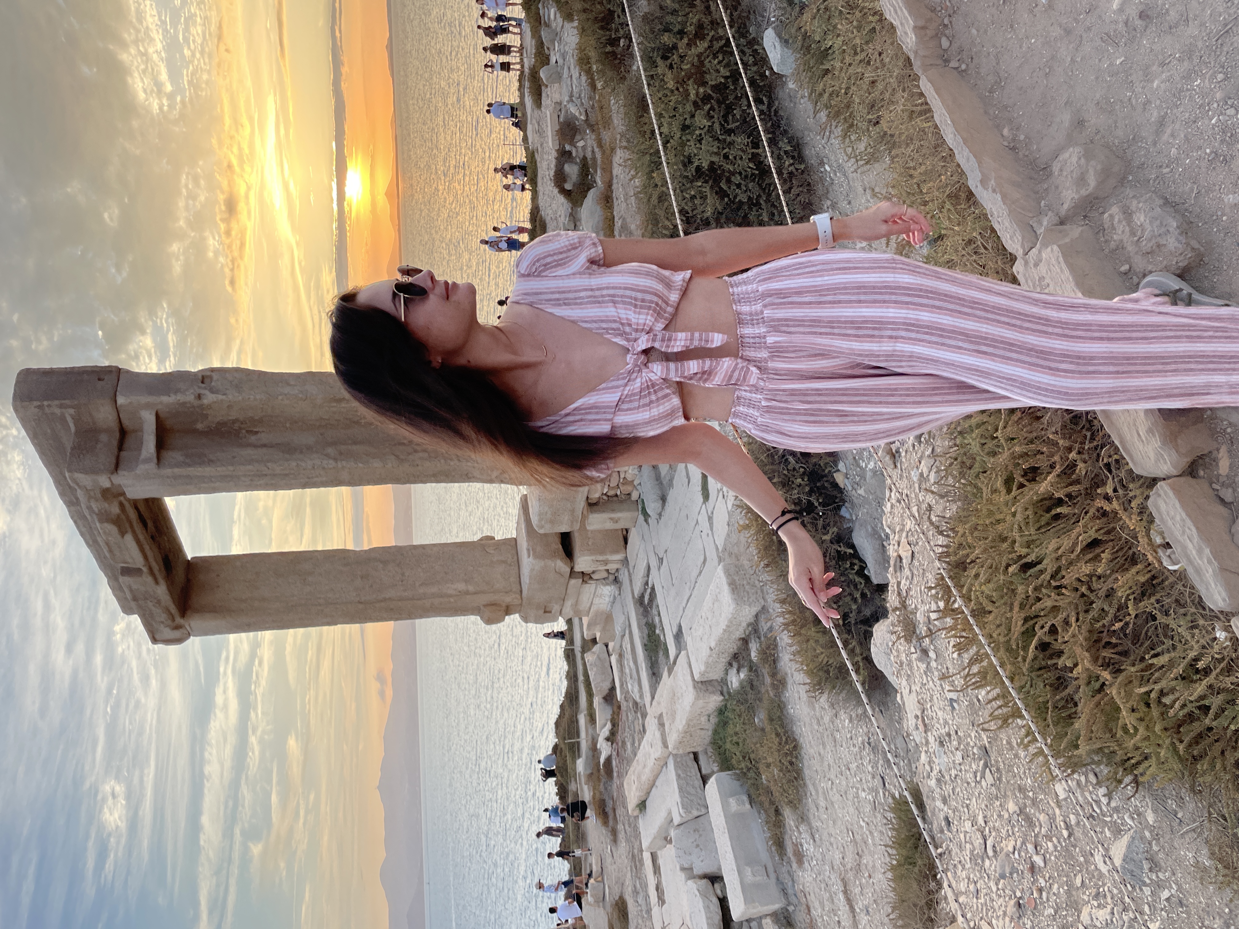 Alayna in a pink and white striped outfit posing in front of an archaeological structure by the sea at sunset