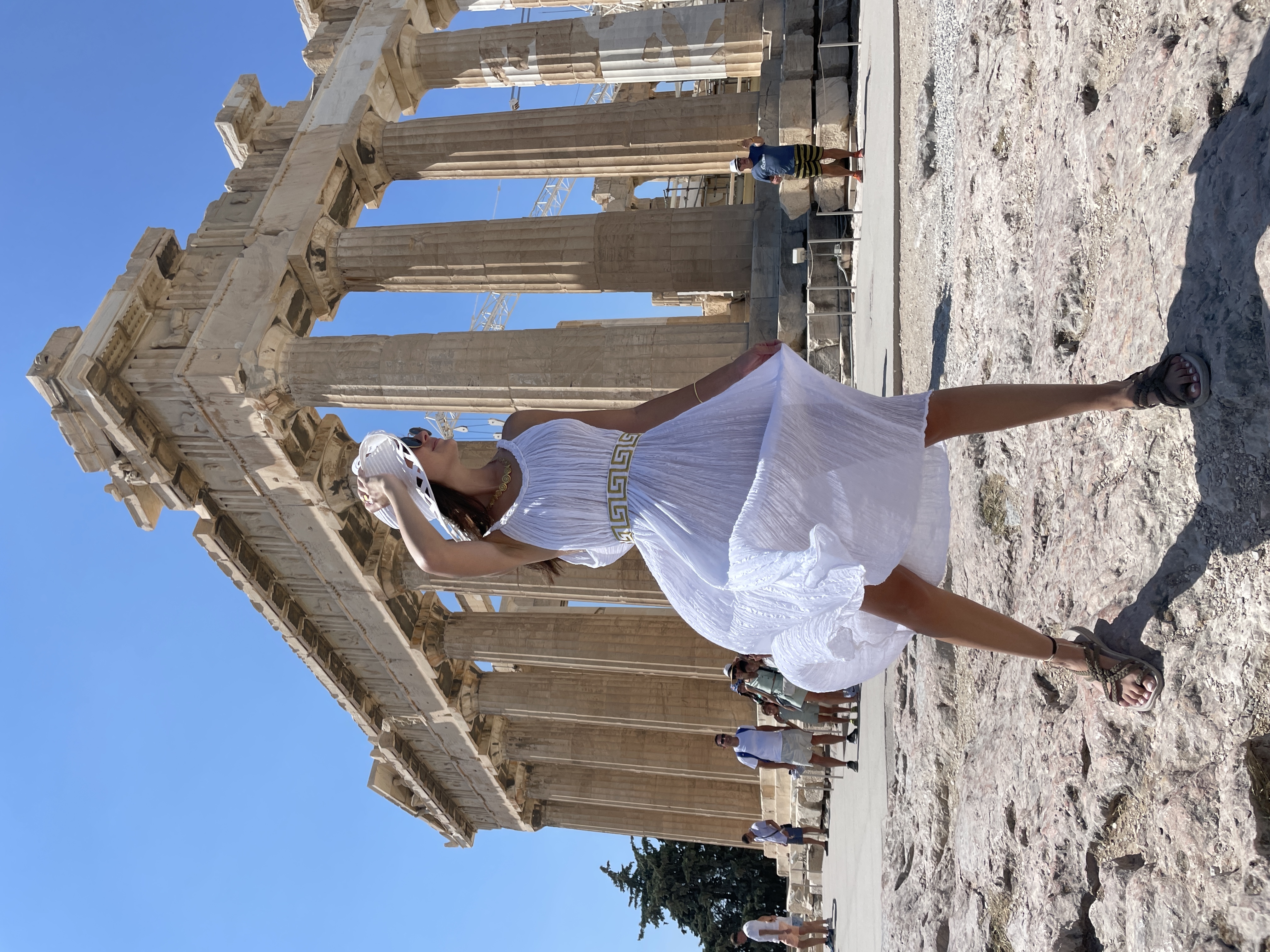 Alayna in a white dress and hat standing in front of the Acropolis on a sunny day