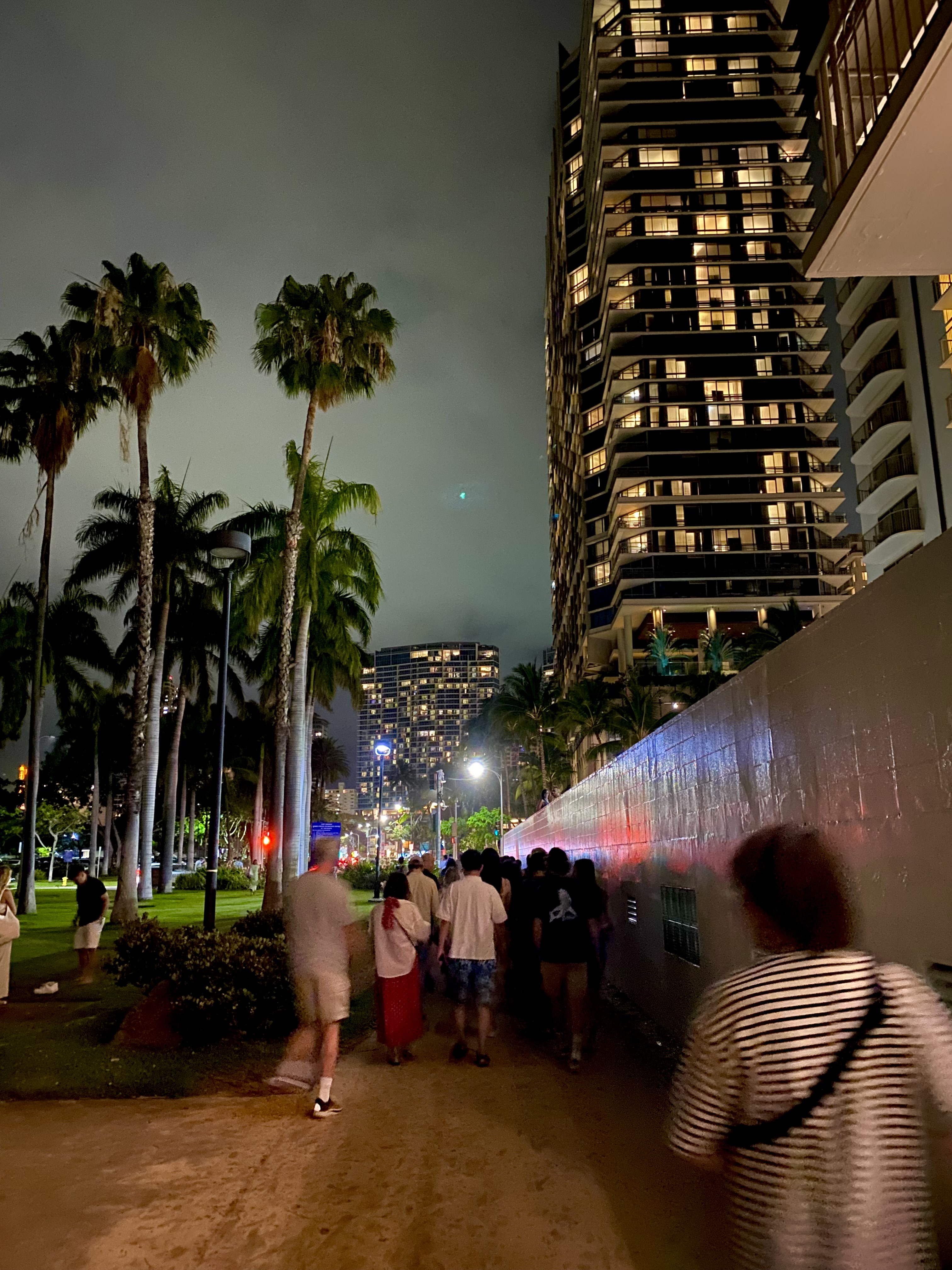 View of pedestrians on a city sidewalk with palm trees and a tall building visible at night