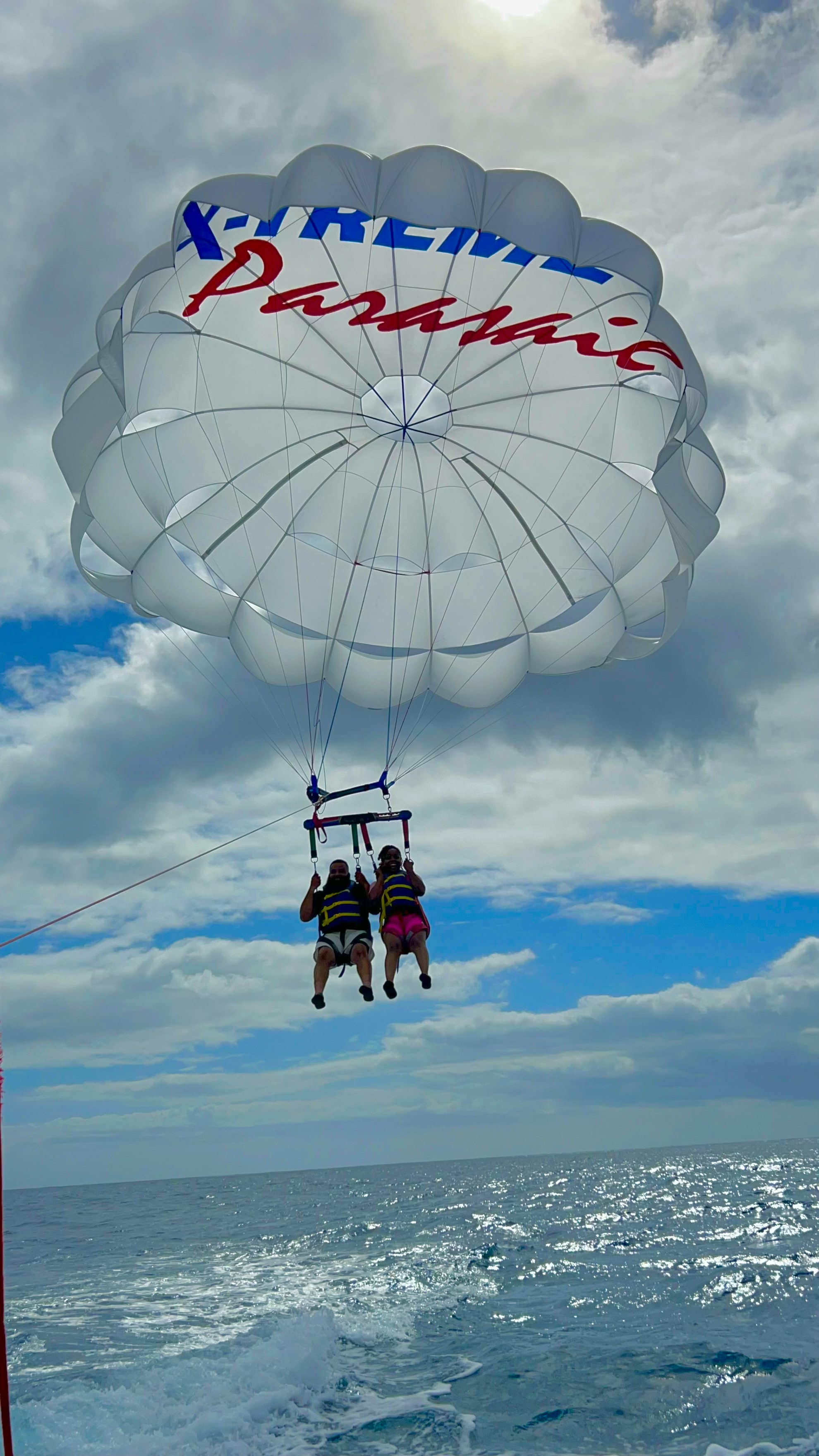 Two people hovering above the ocean with a large white parasail above on a sunny day