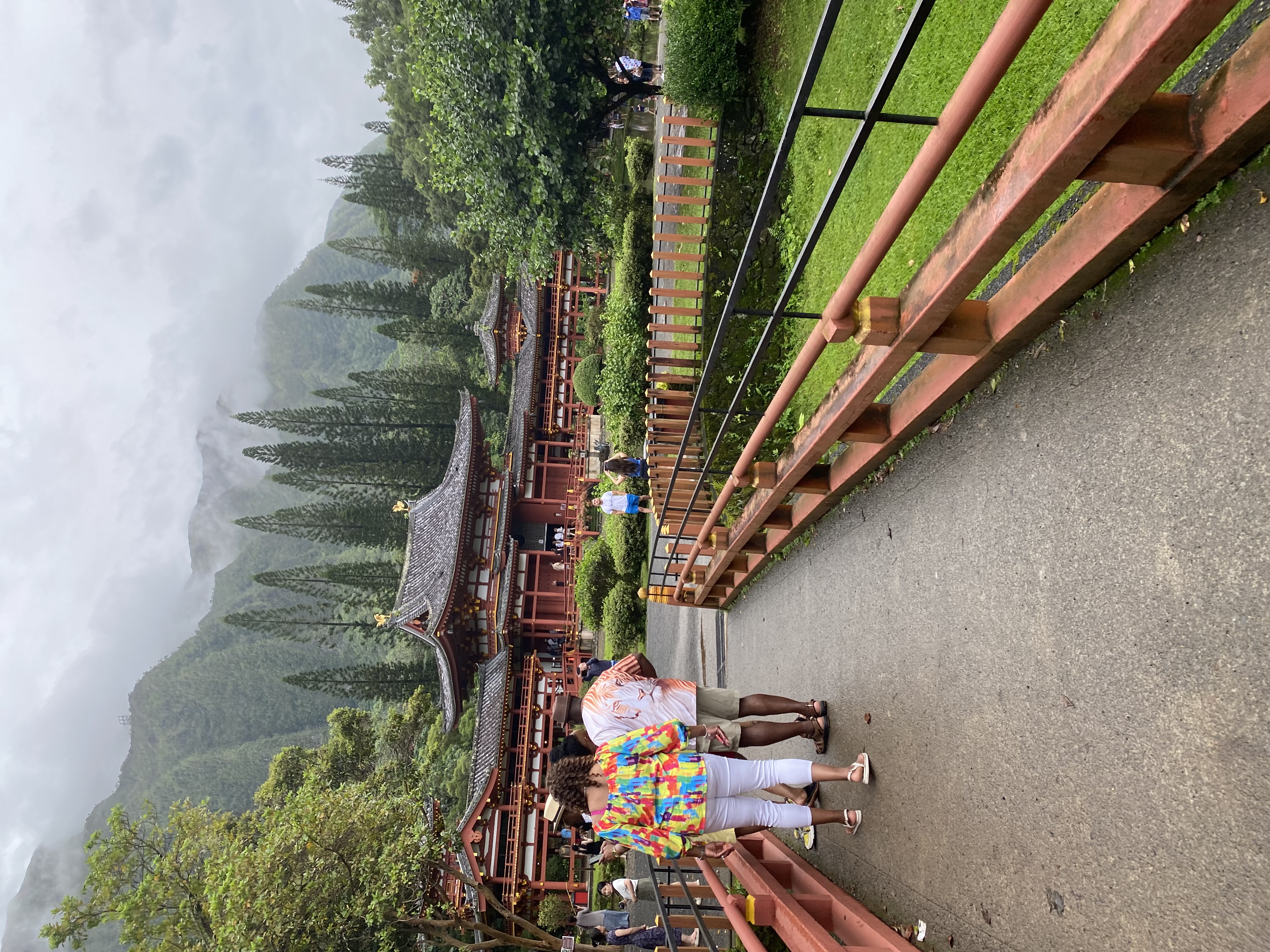 People on a pathway leading towards a large temple with trees and mountains visible behind it on a cloudy day