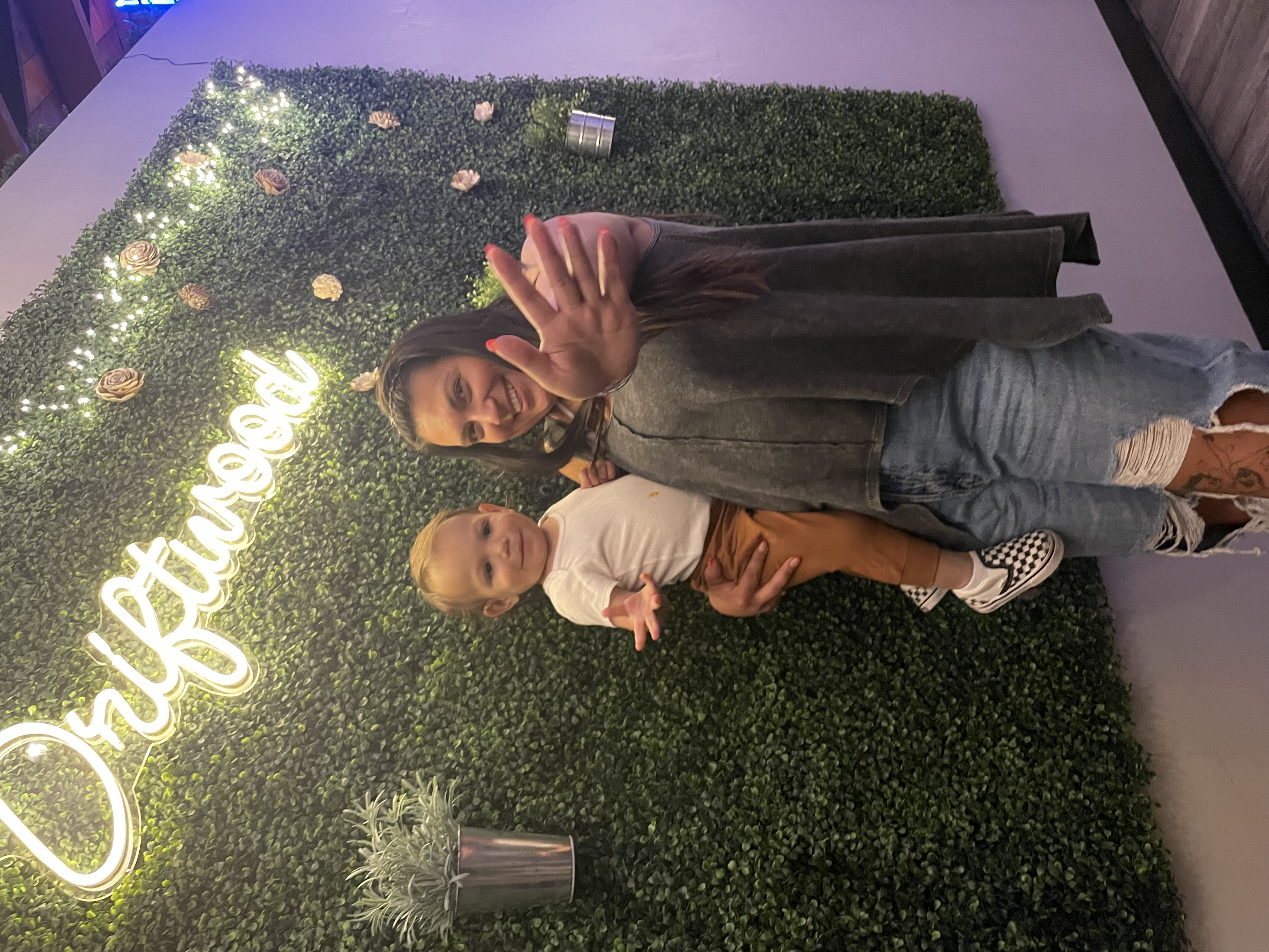 Olivia holding her son in front of a green wall of plants with a neon sign reading “Driftwood”