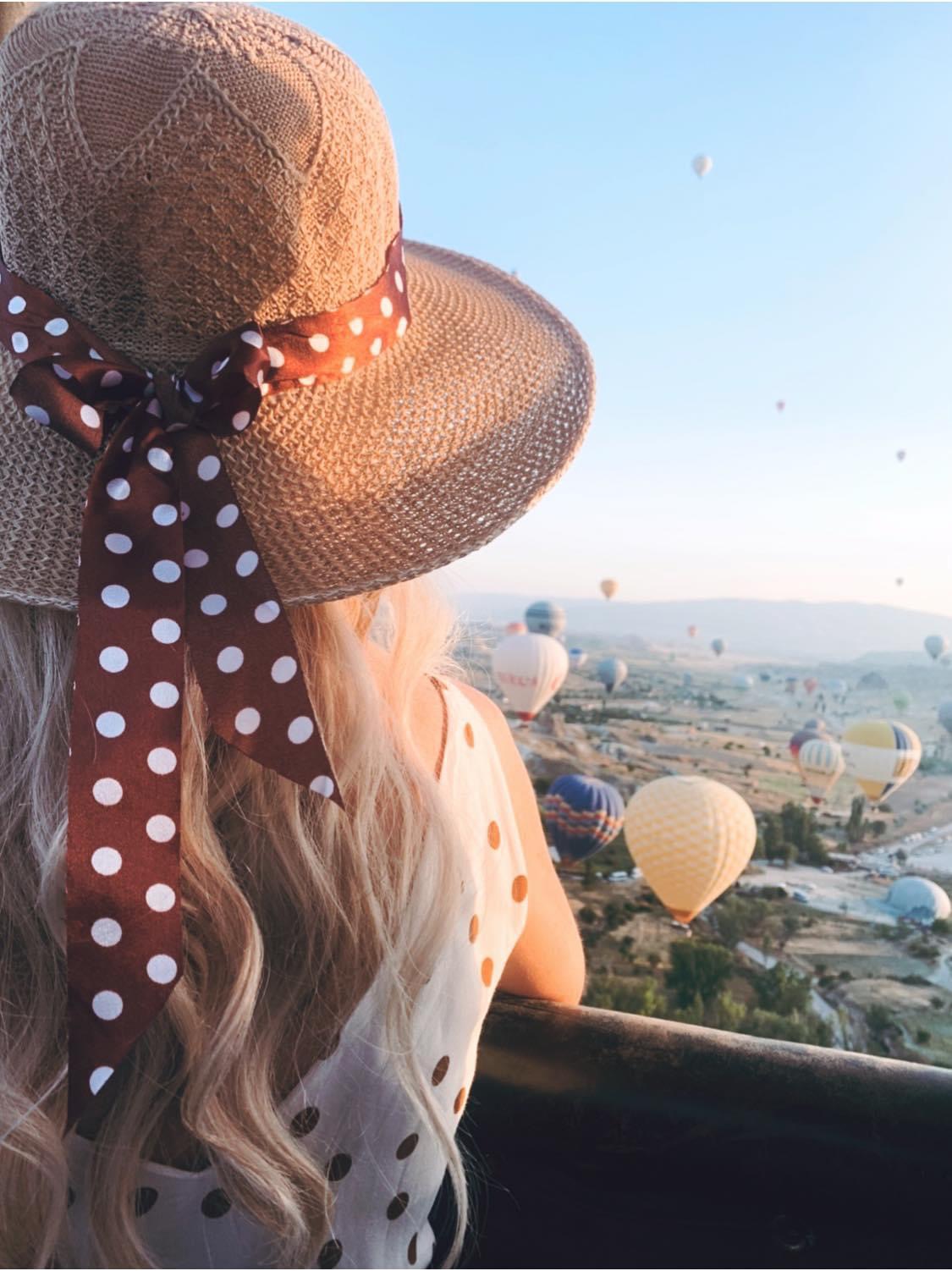 Advisor with long hair, wearing a polka dot hat, looking out at numerous hot air balloons floating in the sky over a scenic landscape.