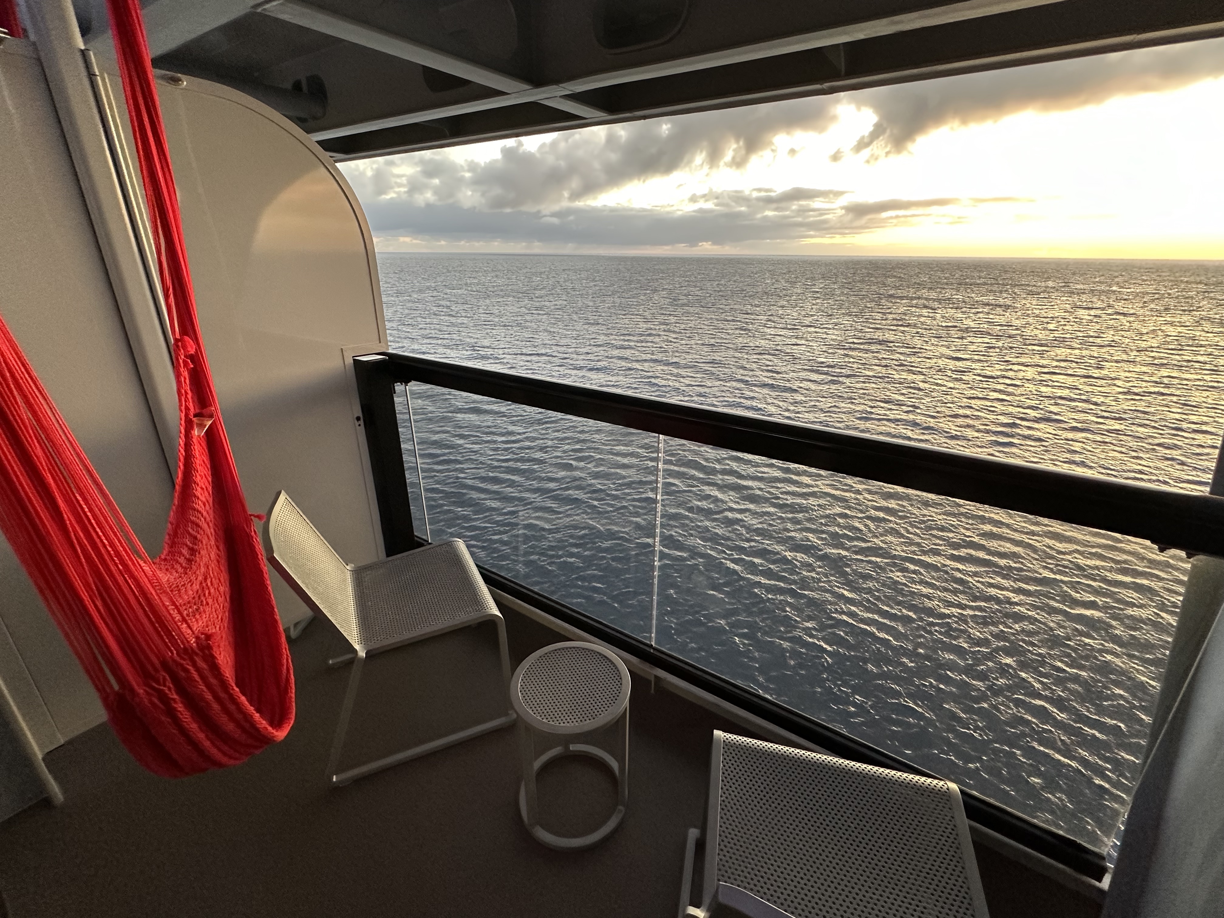 Two chairs and a red hammock on the balcony of a cruise ship overlooking the sea at sunset