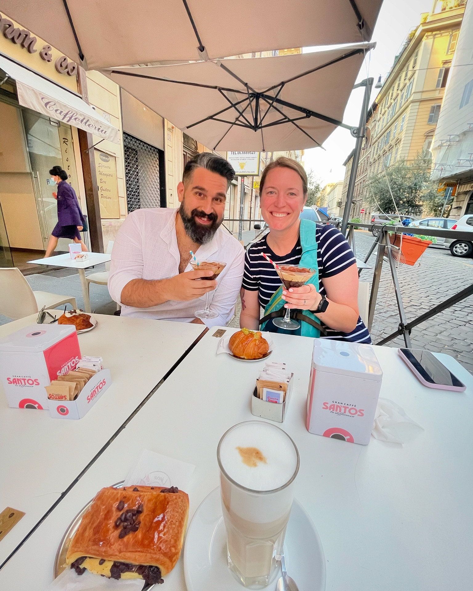 Beth and her partner sitting at an outdoor restaurant table with croissants and coffee in front of them