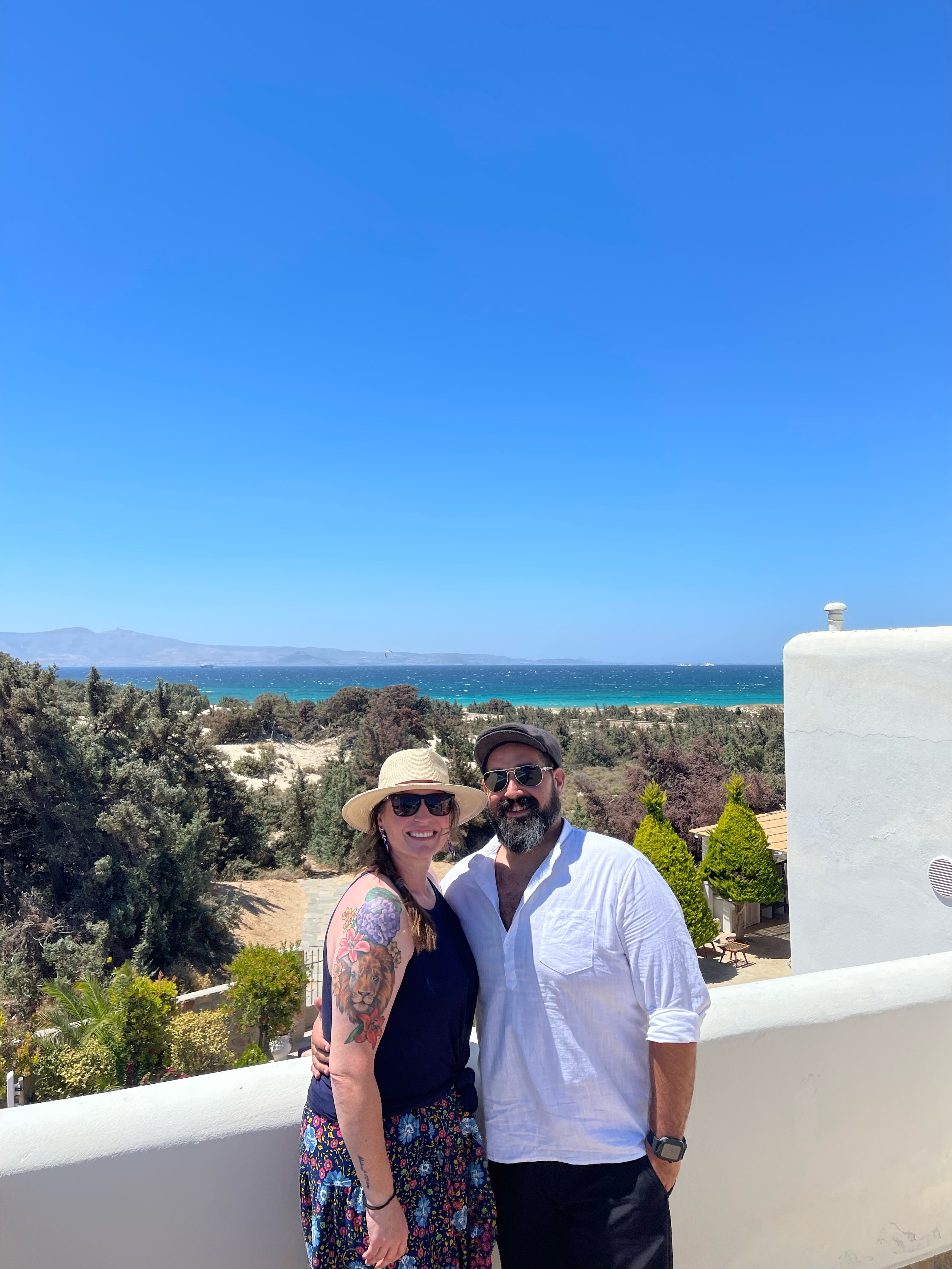 Beth and her partner with arms around each other on a balcony overlooking a beautiful view to the sea under clear skies
