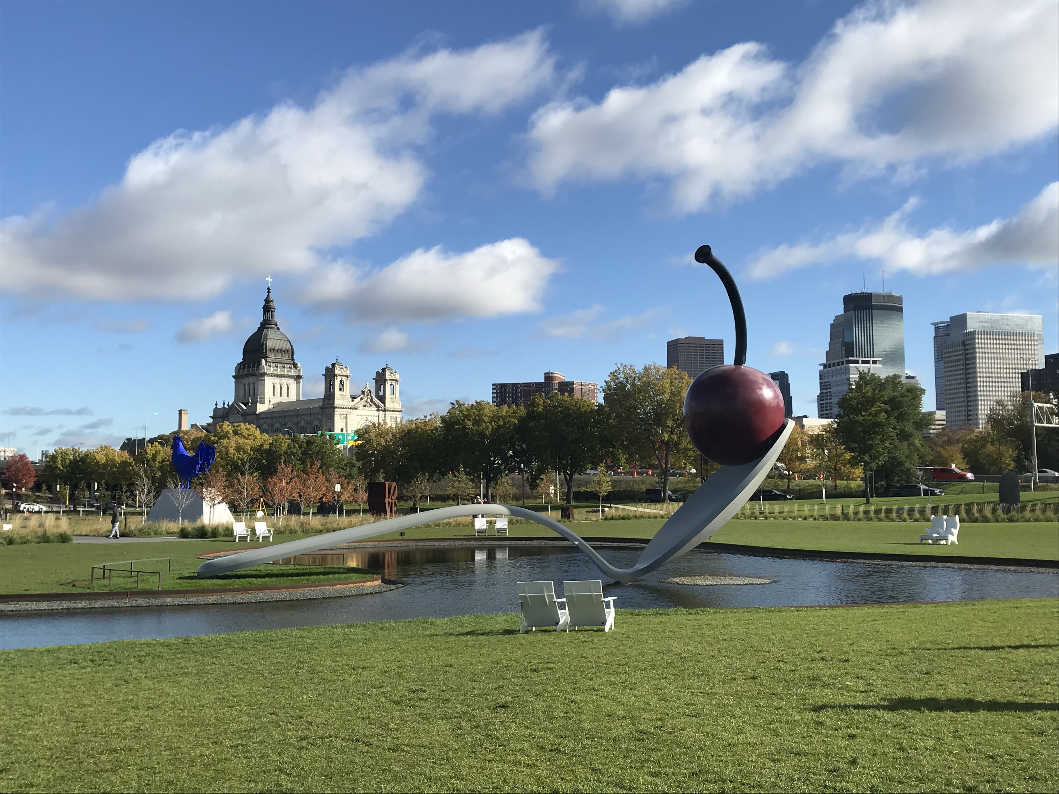 View of a large metal sculpture beside a pond in a green park with city buildings visible behind it