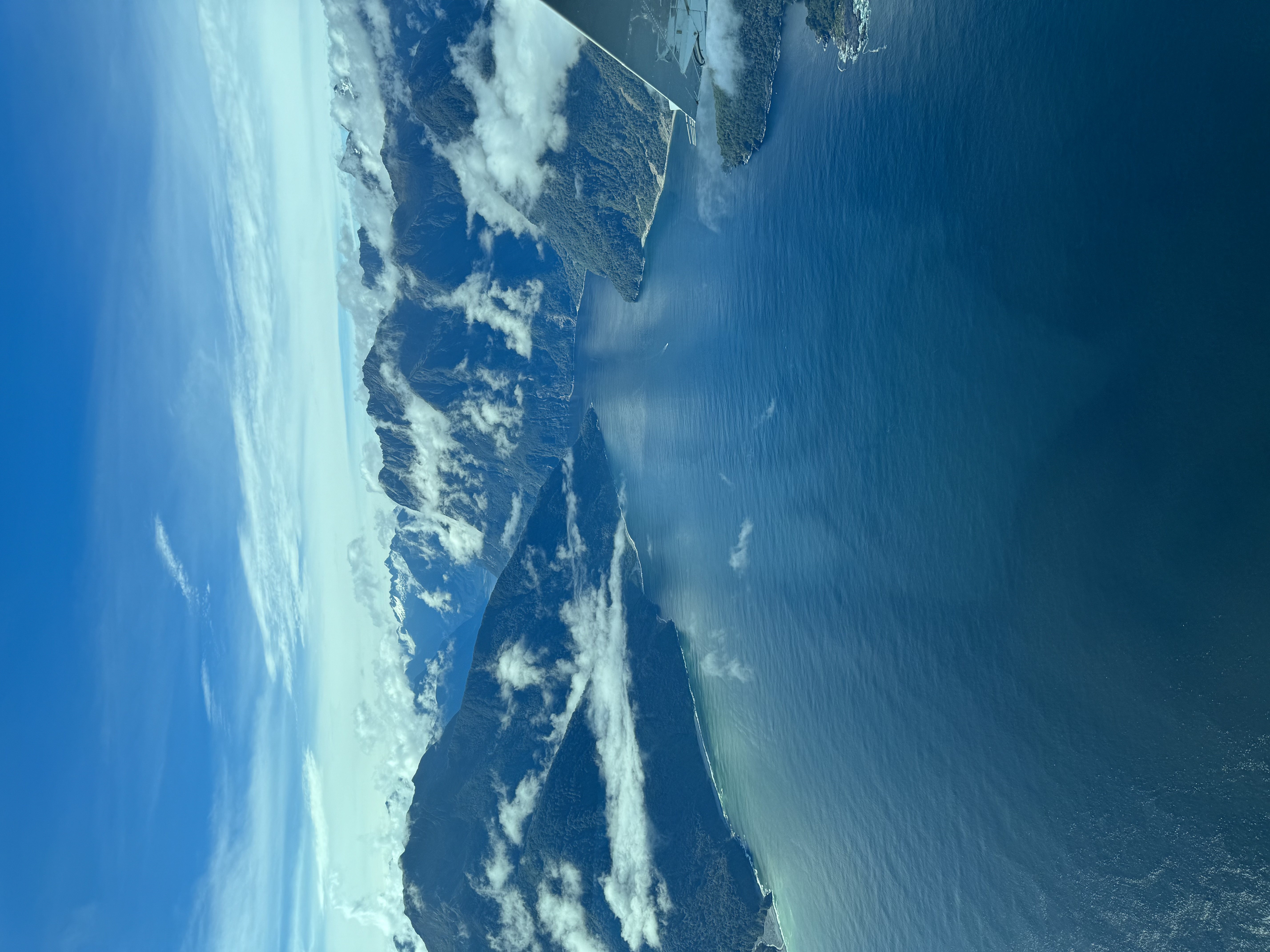 An aerial view of a mountain range with fluffy clouds and a body of water.