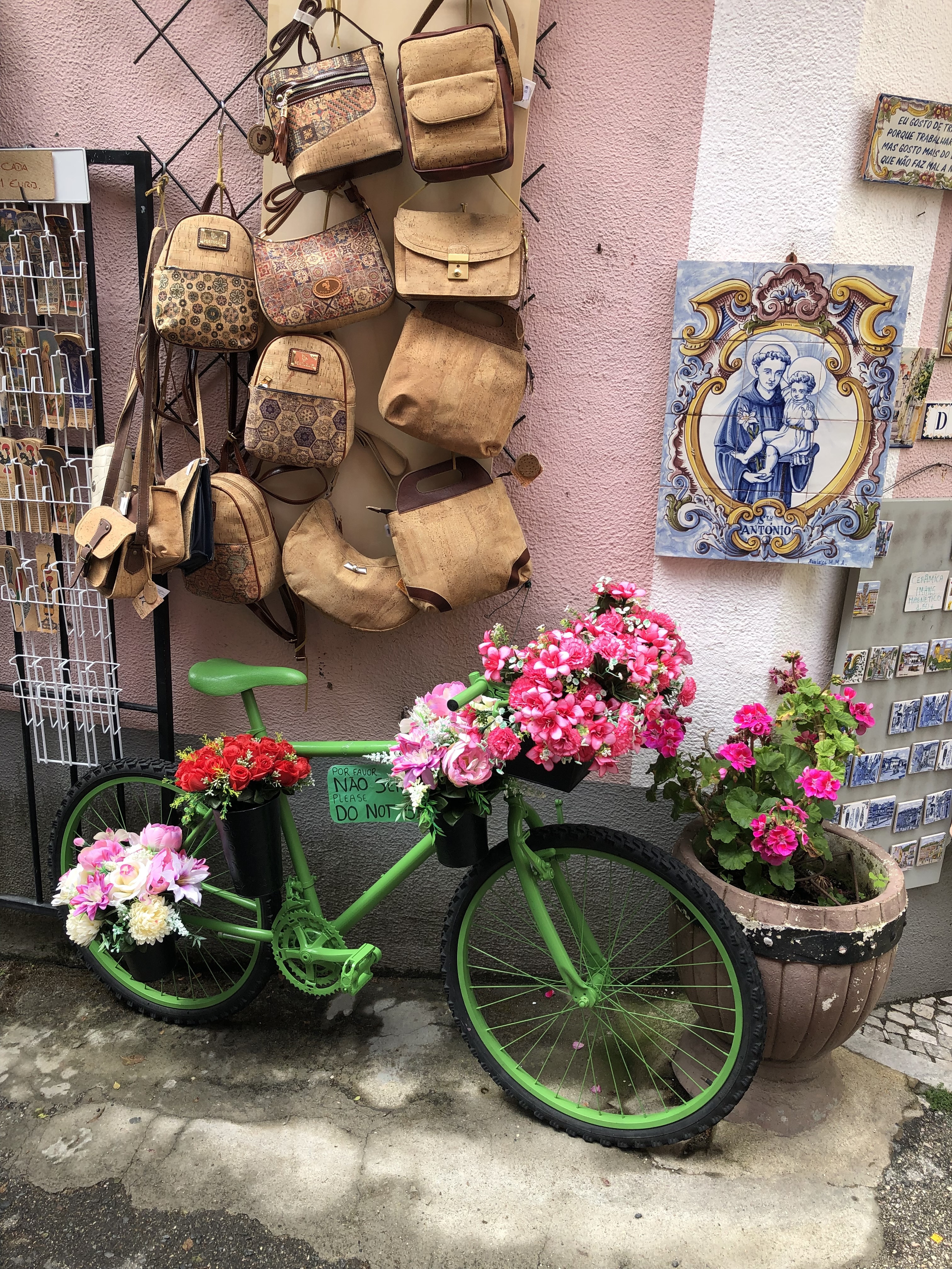 A green bike with pink flowers in a basket parked in front of a wall with bags on display