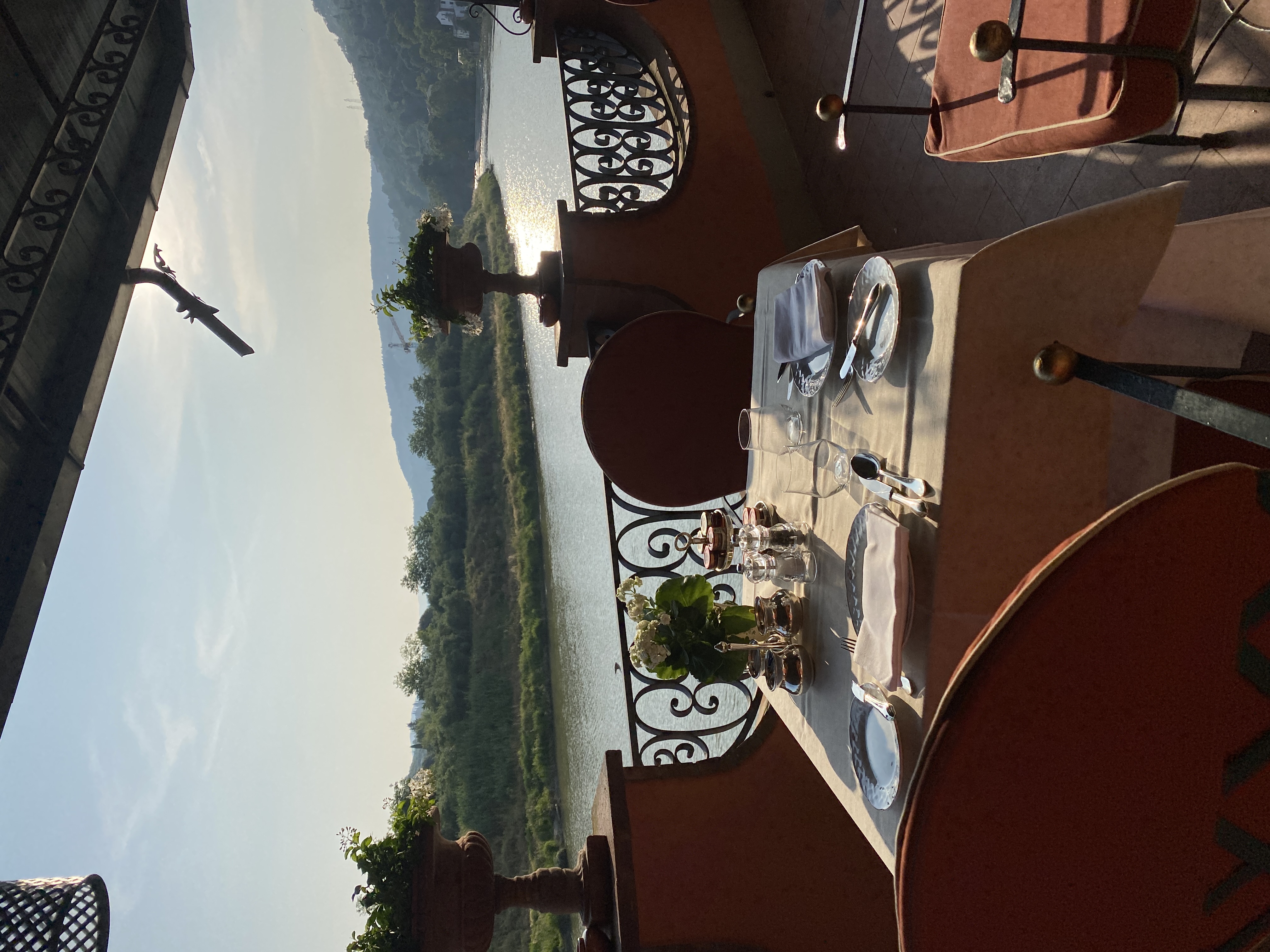 An outdoor restaurant table set for two on a balcony overlooking a beautiful river on a sunny day