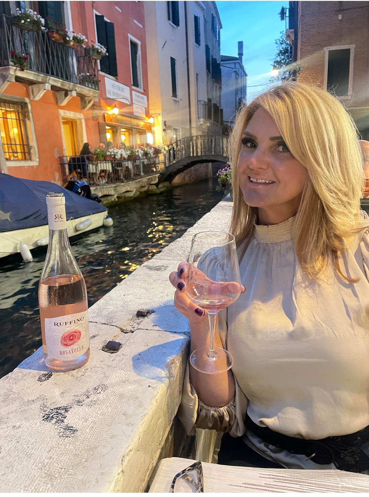 Advisor in a white top seated along a canal in Venice holding a glass of rose wine at sunset