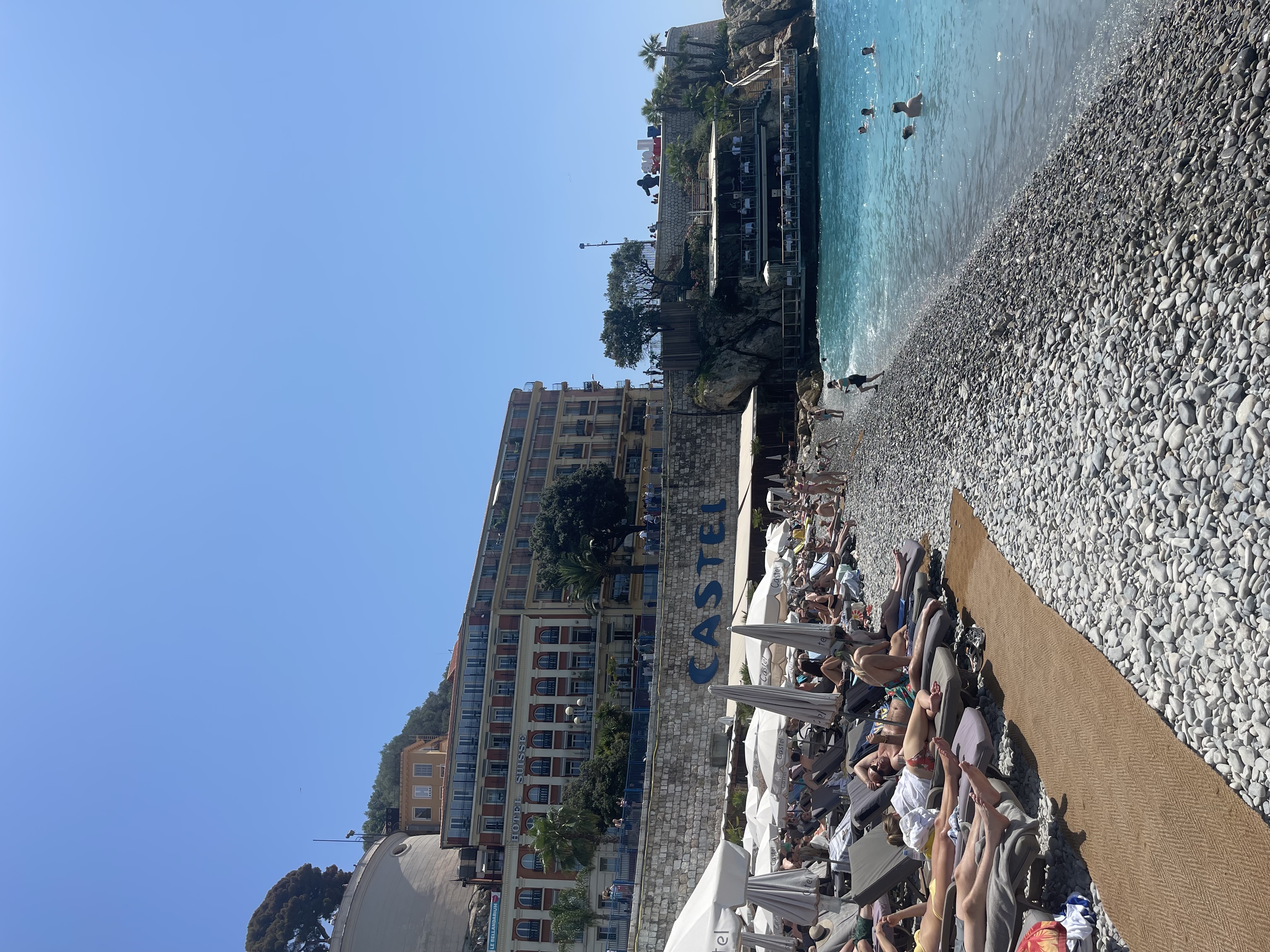 View of a rocky beach and rows of lounge chairs with sunbathers under clear skies