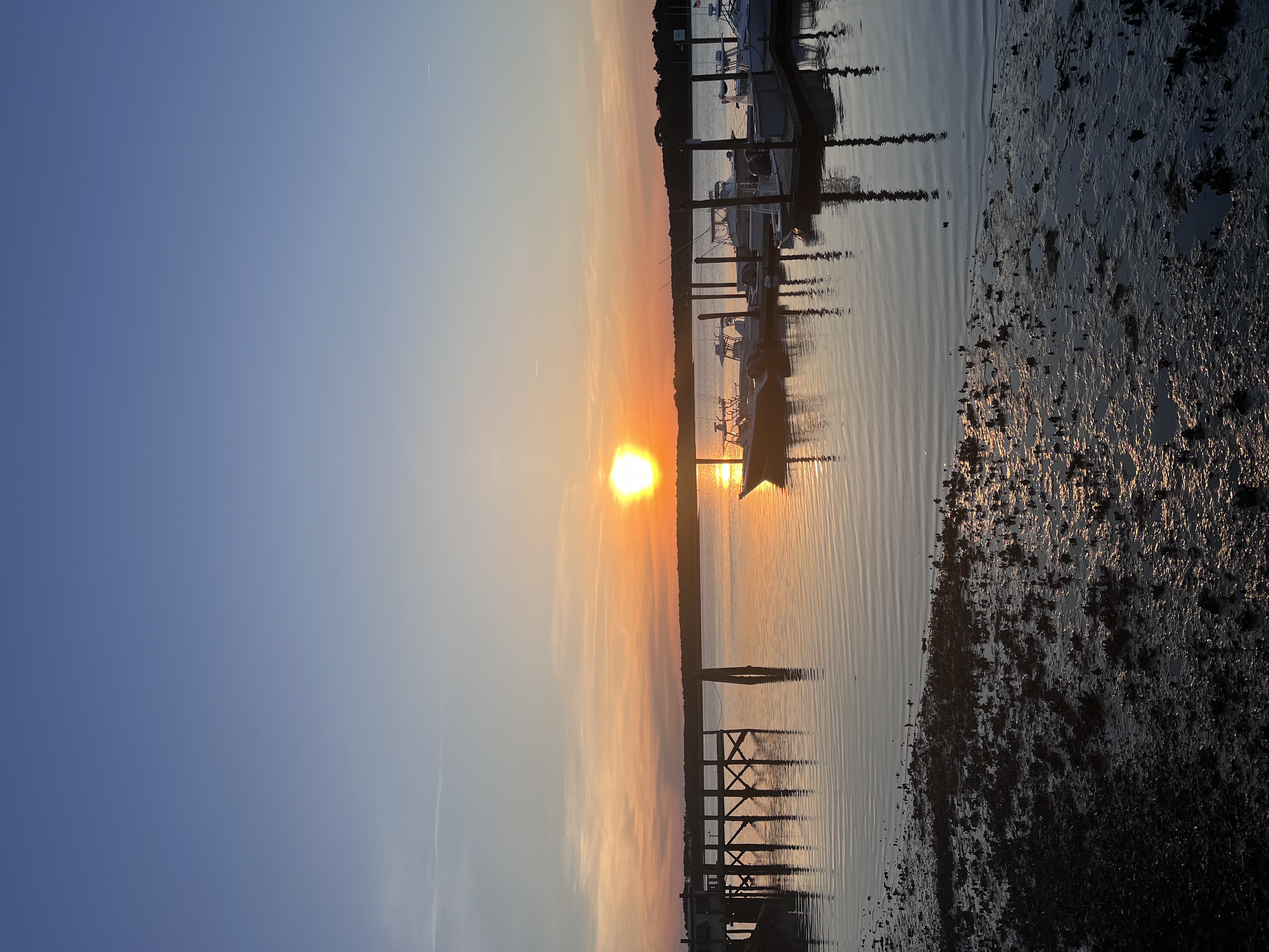 The sun sets over still water near the shore as boats float across the pier on a clear day.