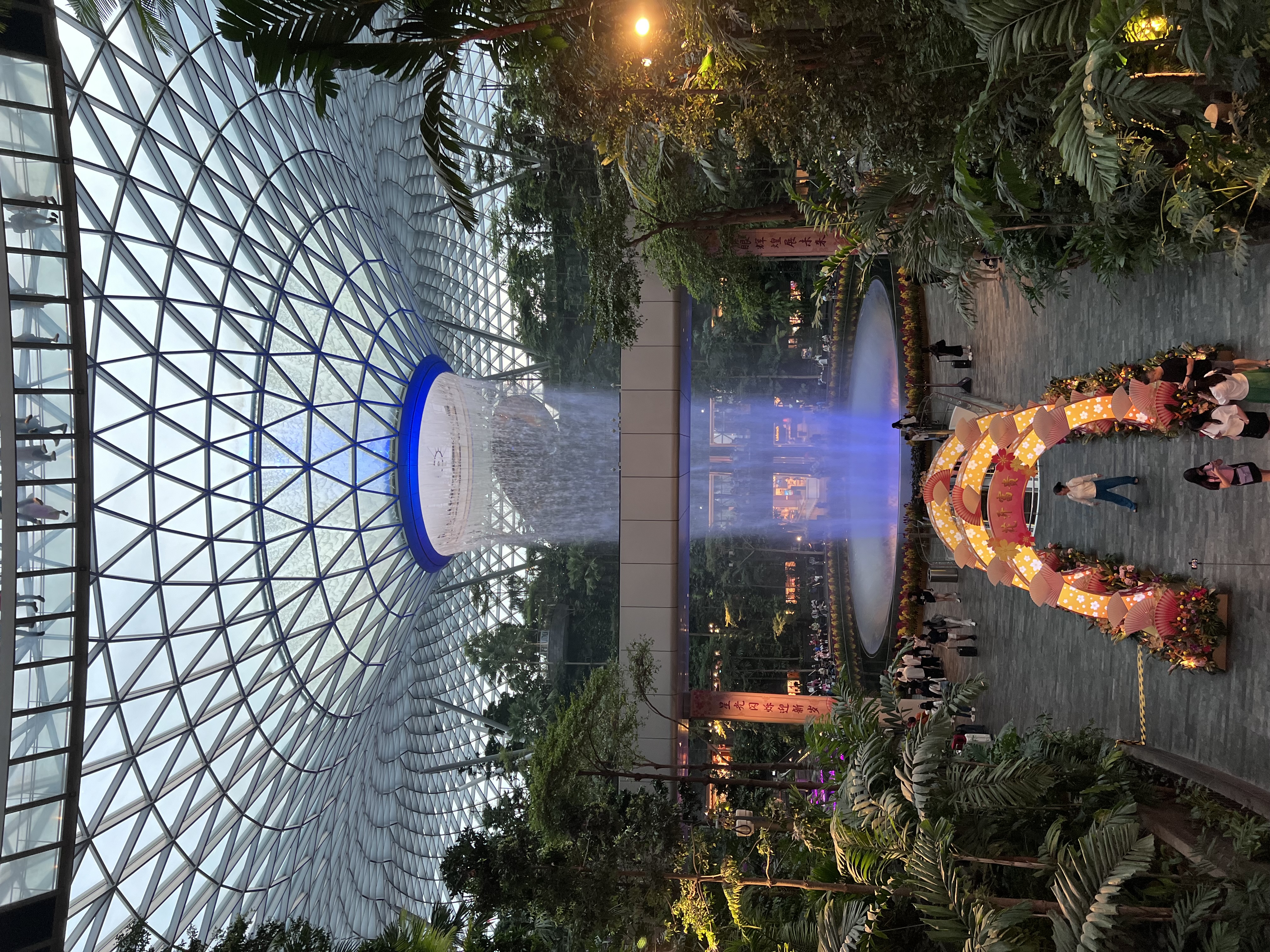 Dense foliage reaches toward the ceiling of an enormous green house lit up on a clear day at dusk.