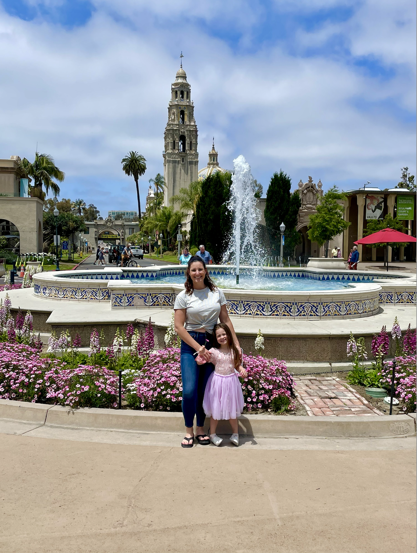 An image of a lady and small child standing in front of a circular fountain with purple flowers surrounding it.