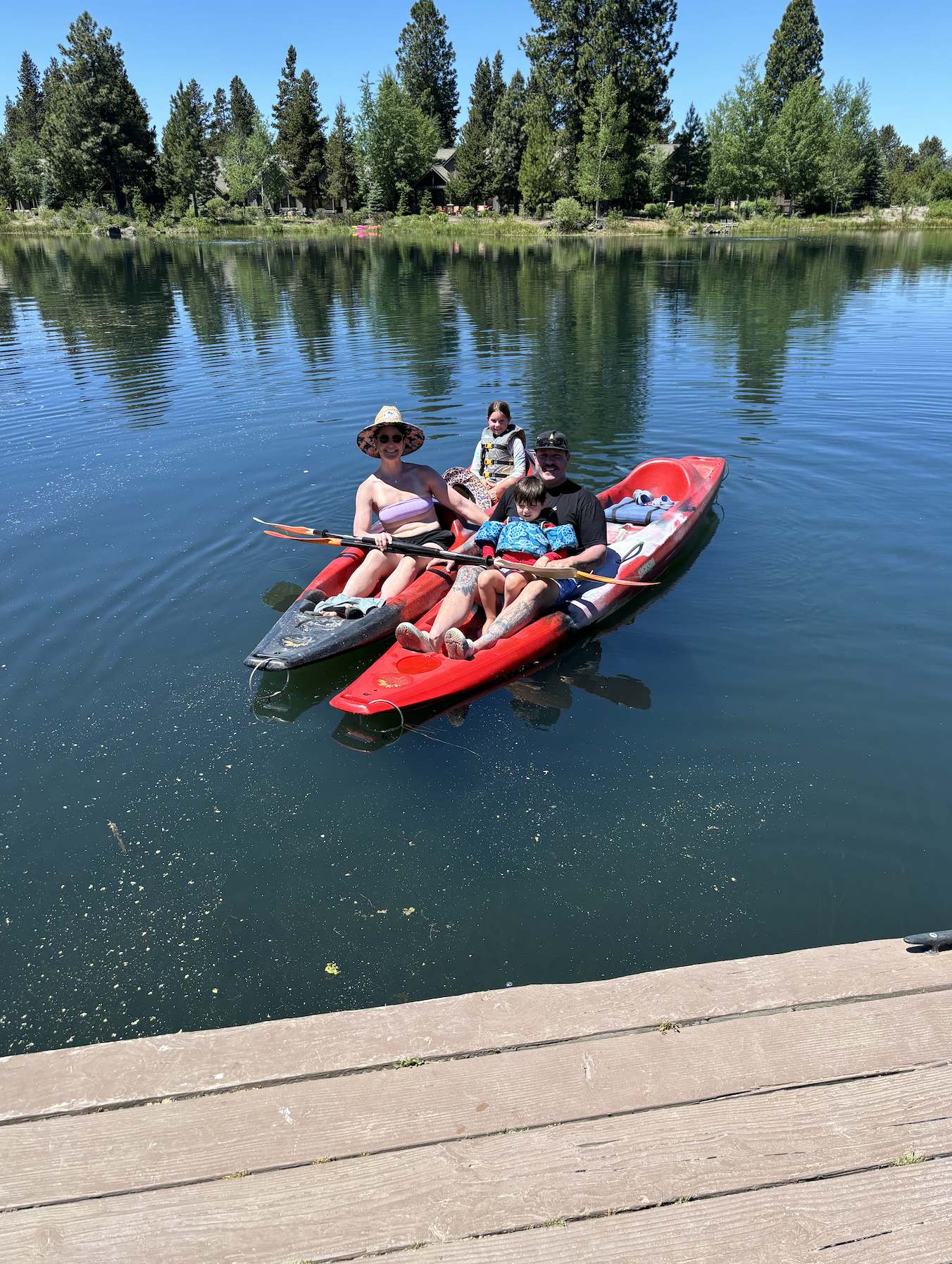 An image of a family of four kayaking on a lake with lush green trees behind.