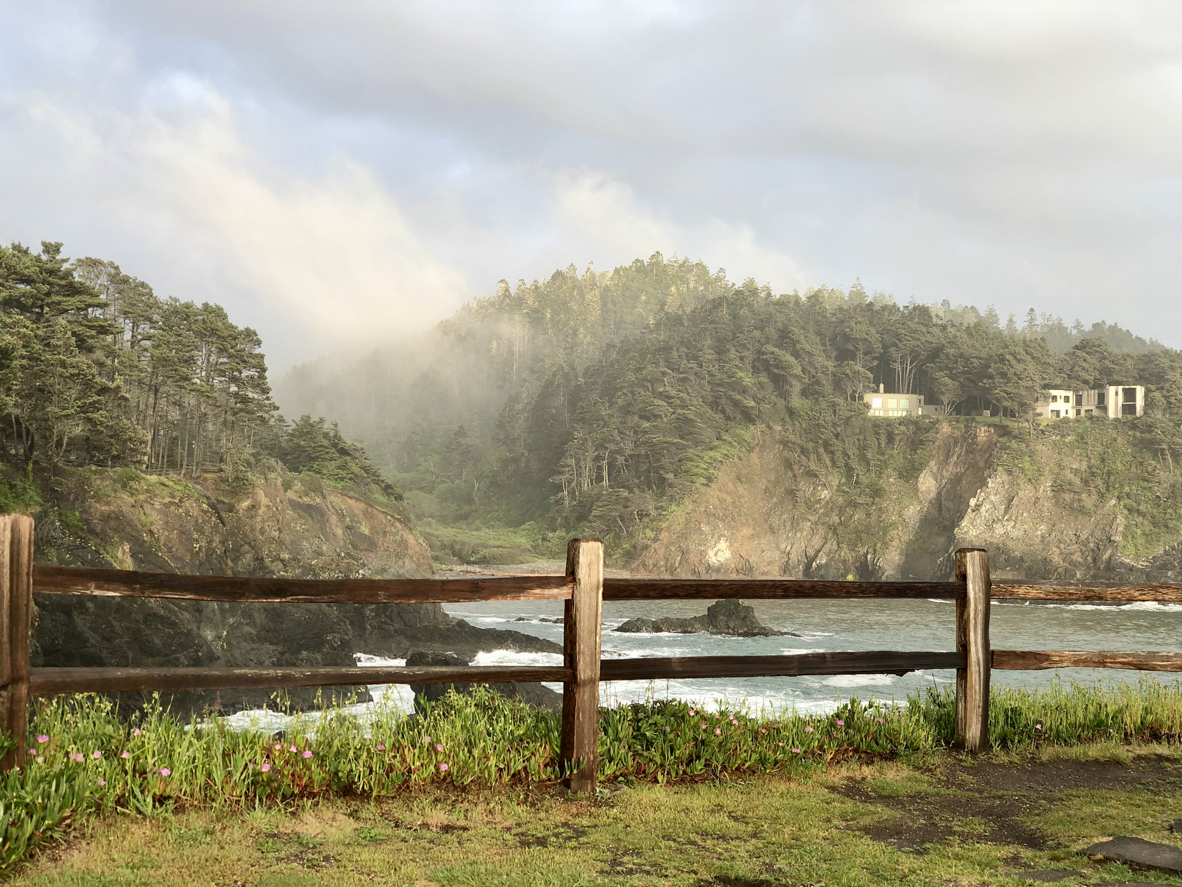 An image of wooden fence in front of the ocean with grassy hills and rocks and a cloudy sky.