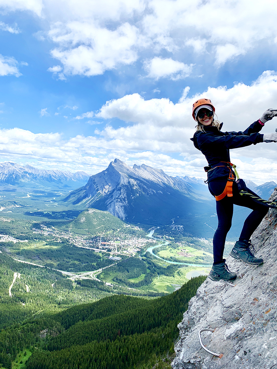 Advisor in rock climbing gear seen on the side of a mountain with a beautiful green valley behind her