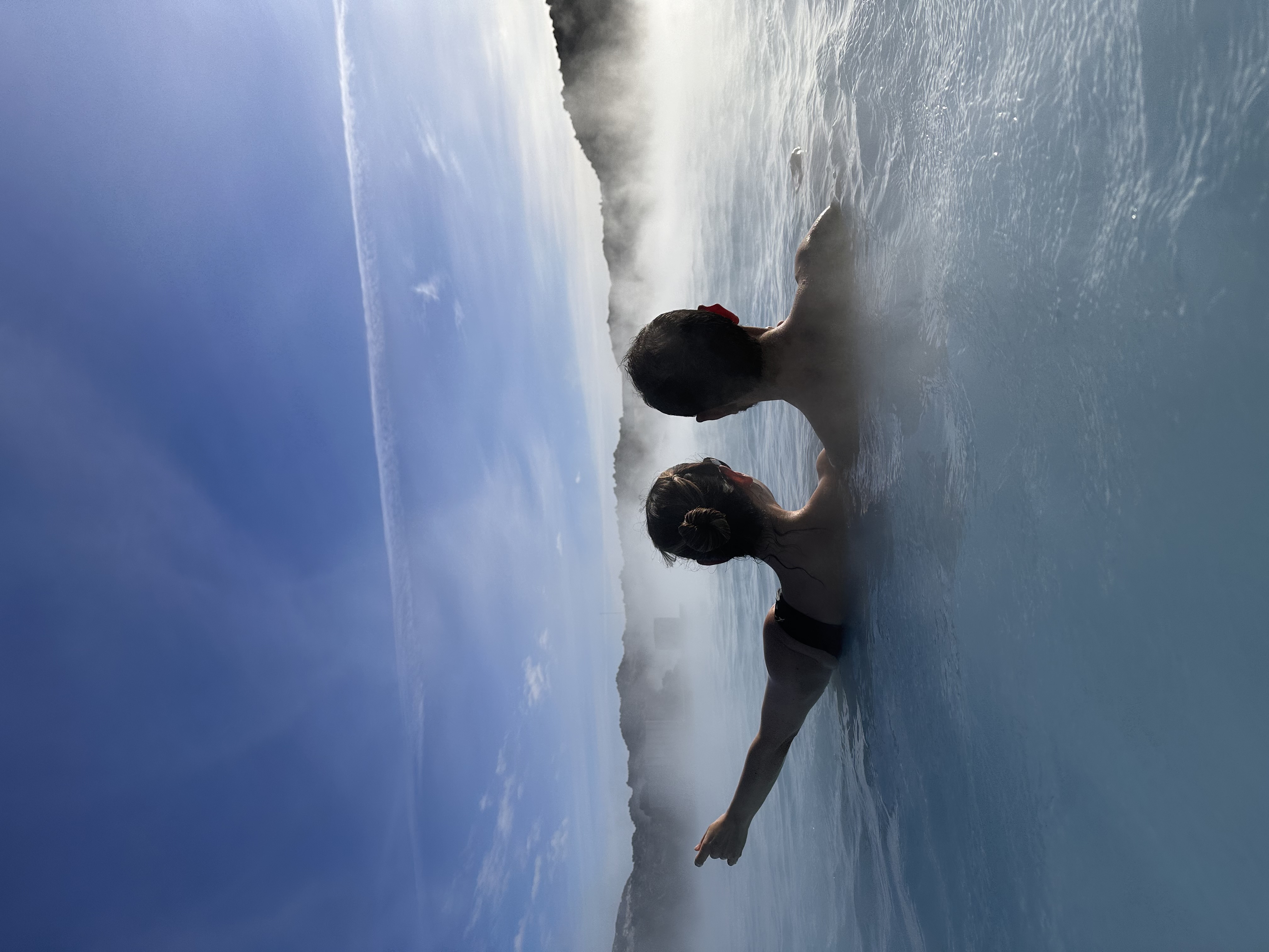 Karla and partner in the water of a geothermal pool. They are viewed from behind, with one person pointing towards something in the distance.