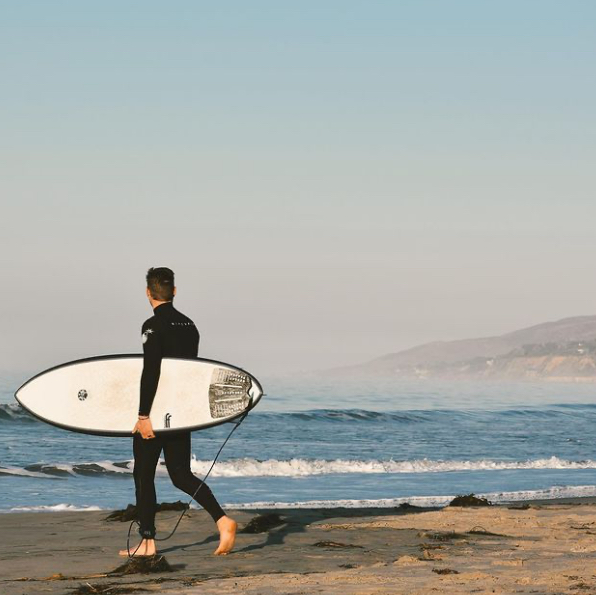 Man in a wetsuit carrying a white surfboard on the beach