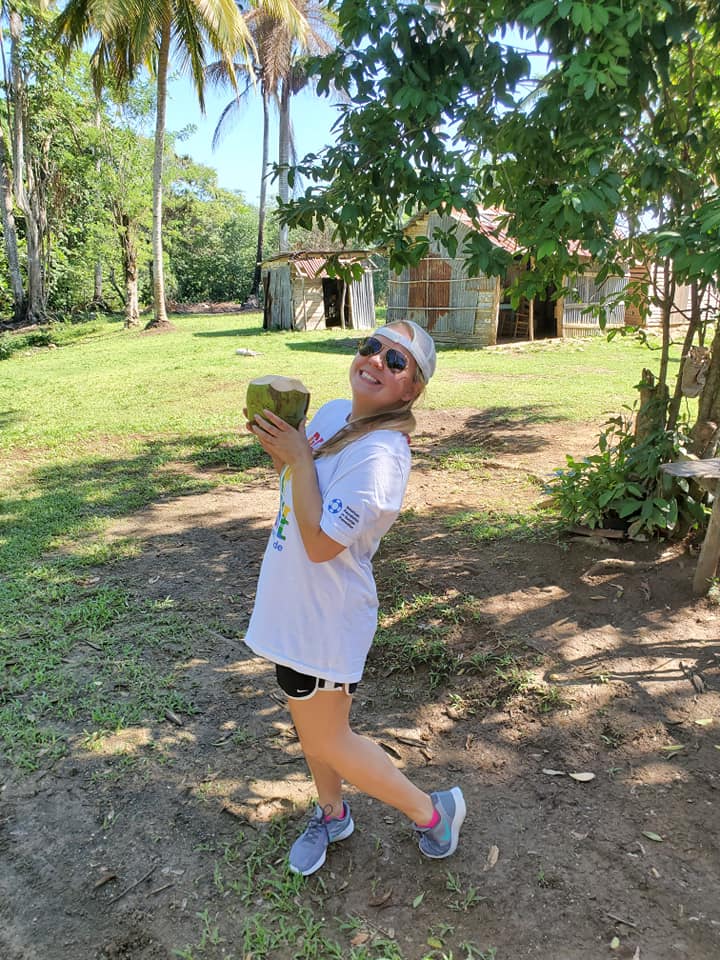A person holding a coconut outside in nature, surrounded by trees and a small shack in the background.