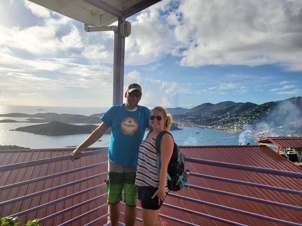A couple posing in an overlook with a view of mountainous towns and a body of water in the background.