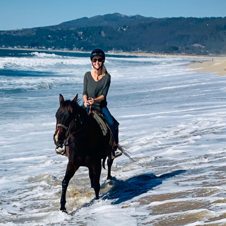 Advisor on a black horse riding through shallow waves on the beach with mountains in the distance