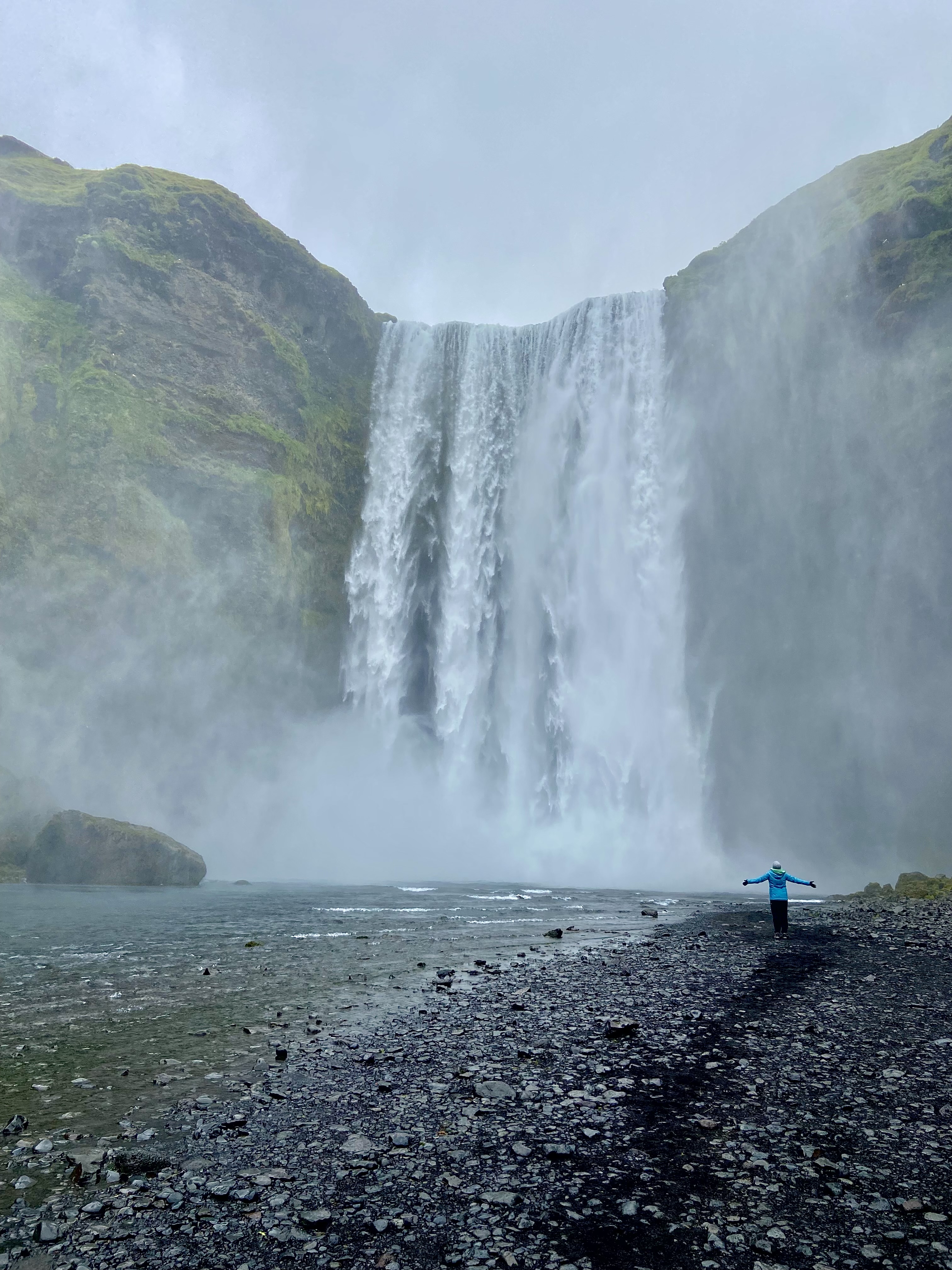 A large waterfall rapidly bellowing downward while a rocky beach and person wearing a blue jacket with their arms outstretched wide awaits at the bottom.